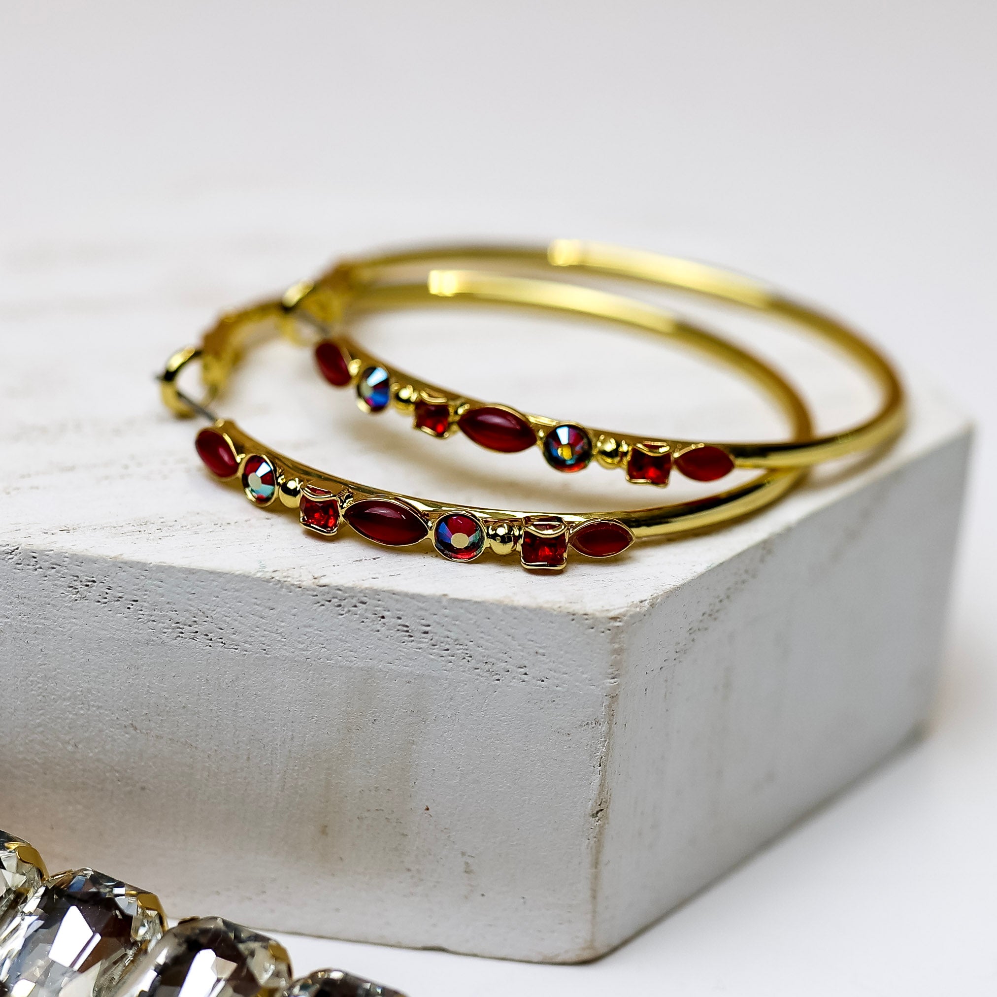 A pair of gold-tone circle hoop earrings with dark red crystals on the front. Pictured on a white background with crystal necklaces.