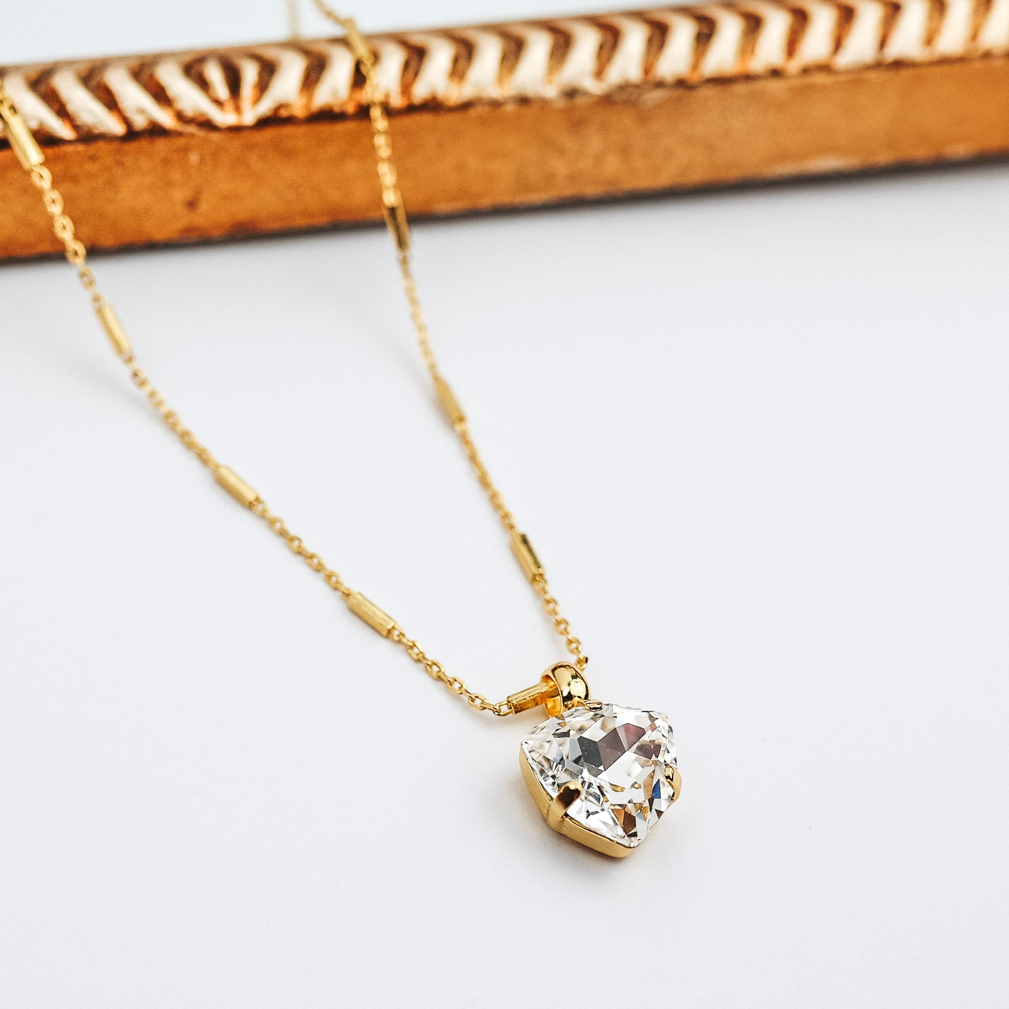 A gold-tone chain necklace with a triangular cut crystal pendant. This necklace is pictured on a white background with a mirror.