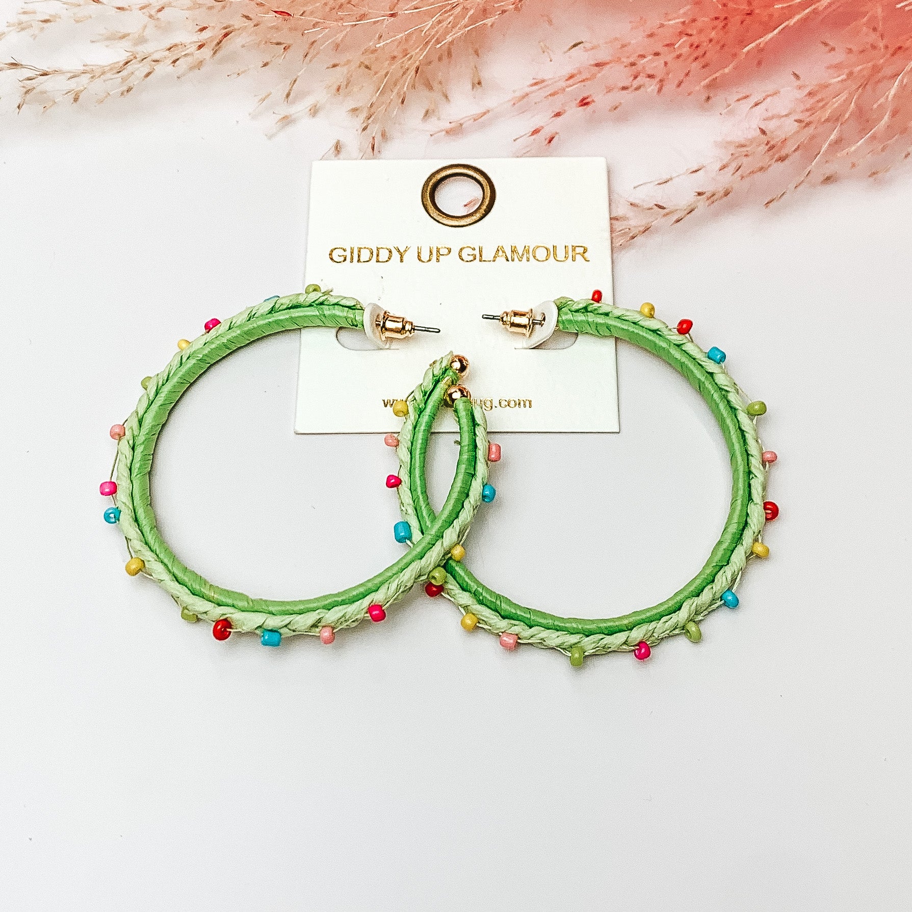 Pictured are raffia braided hoop earrings in light green with colorful beads. They are pictured with a pink feather on a white background.