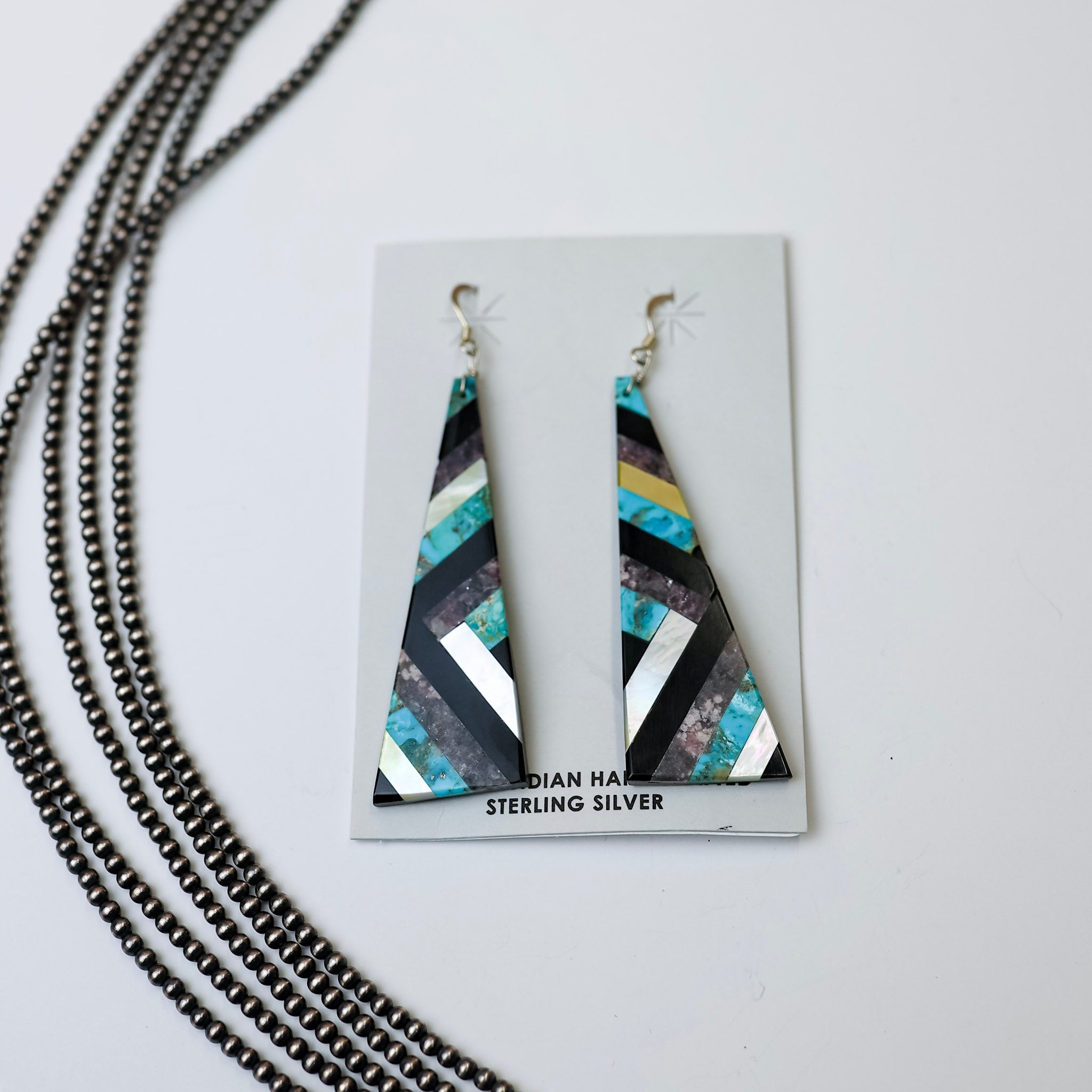 Navajo Handmade Turquoise, Opal, and Onyx Geometric Design Slab Earrings are centered in the middle of the picture. On the left side, Navajo pearls are laid. All on a white background.  
