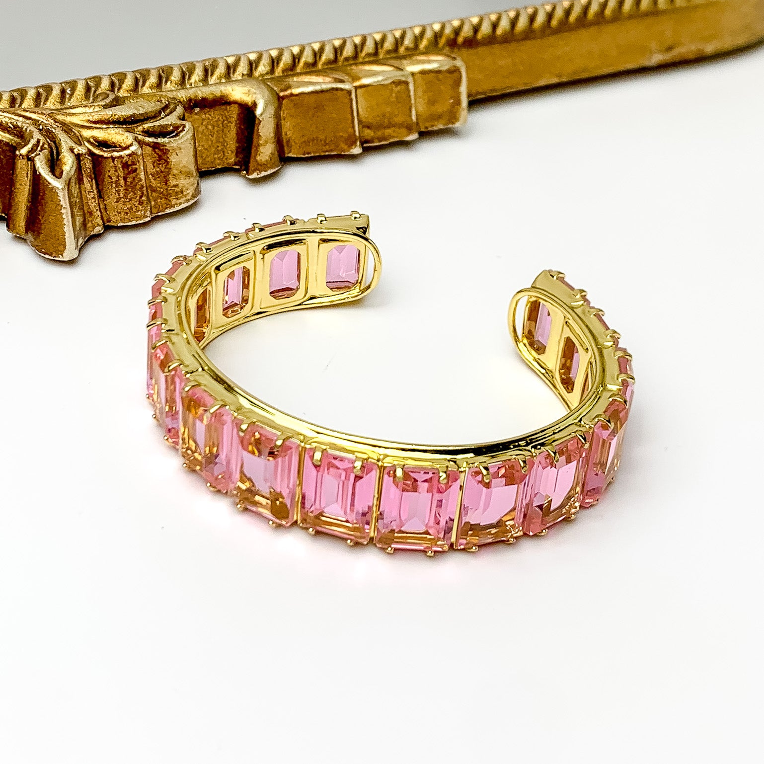 Gold bangle with a pink, rectangle crystal inlay. This bracelet is pictured on a white background with a gold mirror at the top of the picture.  