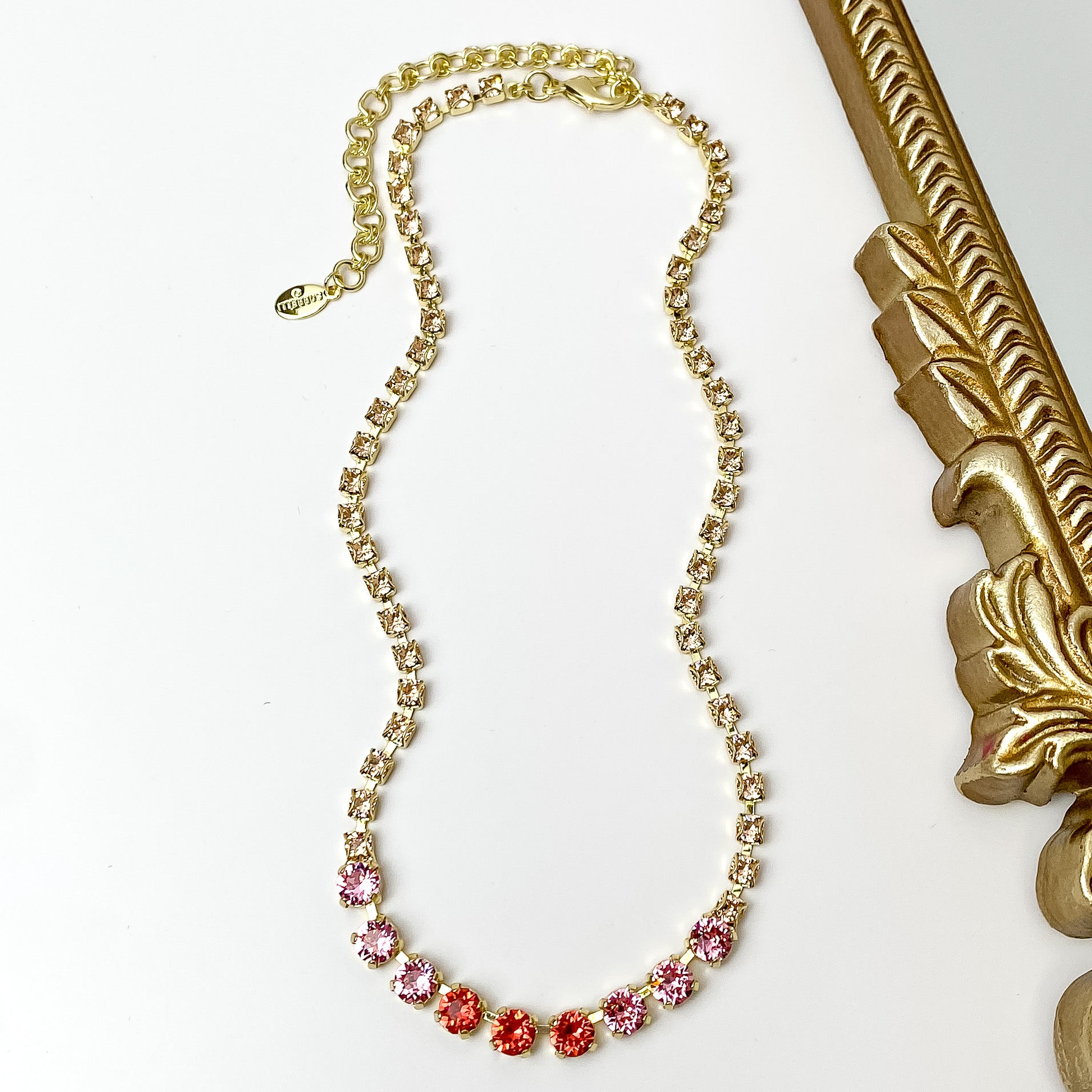 Pictured is a gold necklace with a mix of pink crystals. This necklace is pictured on a white background with a gold mirror on the right side.