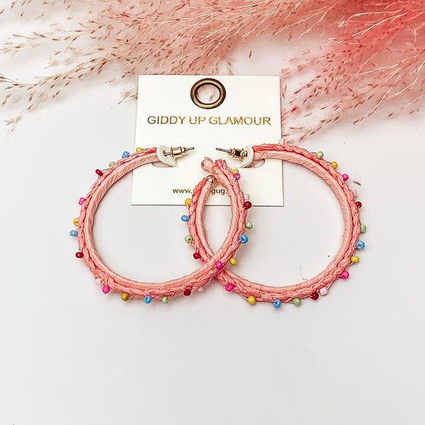 Pictured are raffia braided hoop earrings in light pink with colorful beads. They are pictured with a pink feather on a white background.