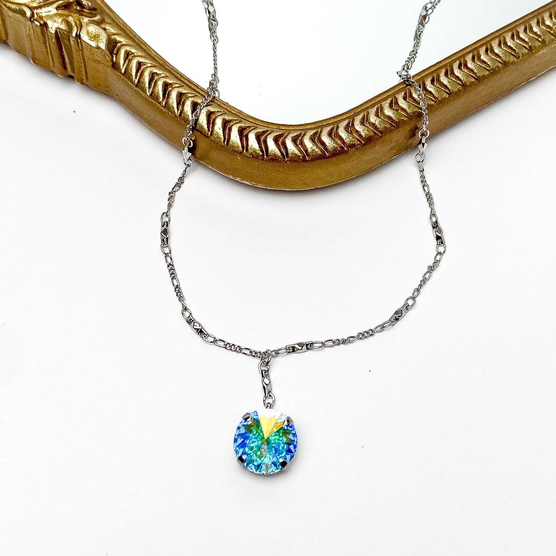 Pictured is a silver necklace with a ab crystal pendant. This necklace is pictured laying partially on a gold mirror on a white background. 