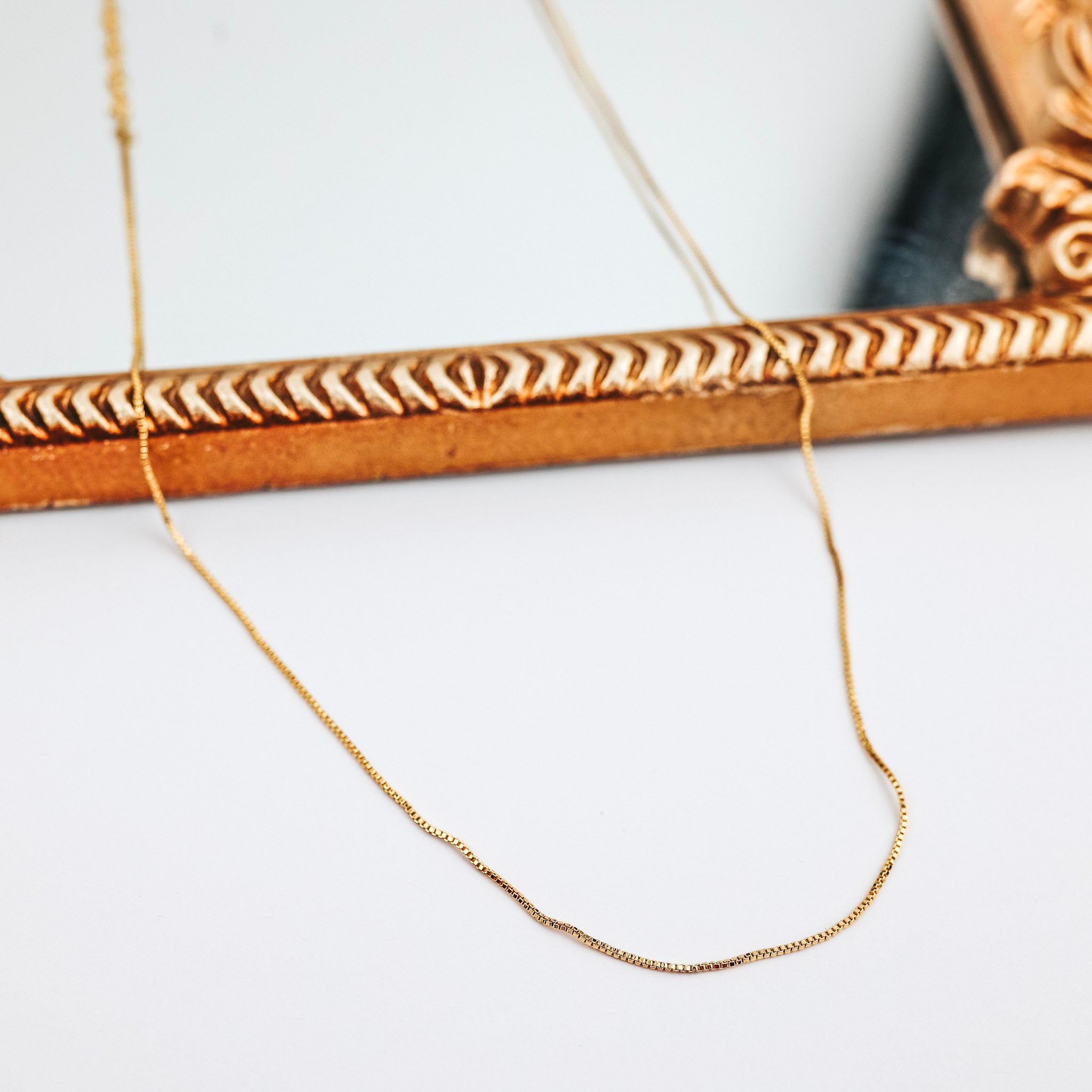 A gold-tone thin box chain necklace that is pictured on a white background with a mirror.