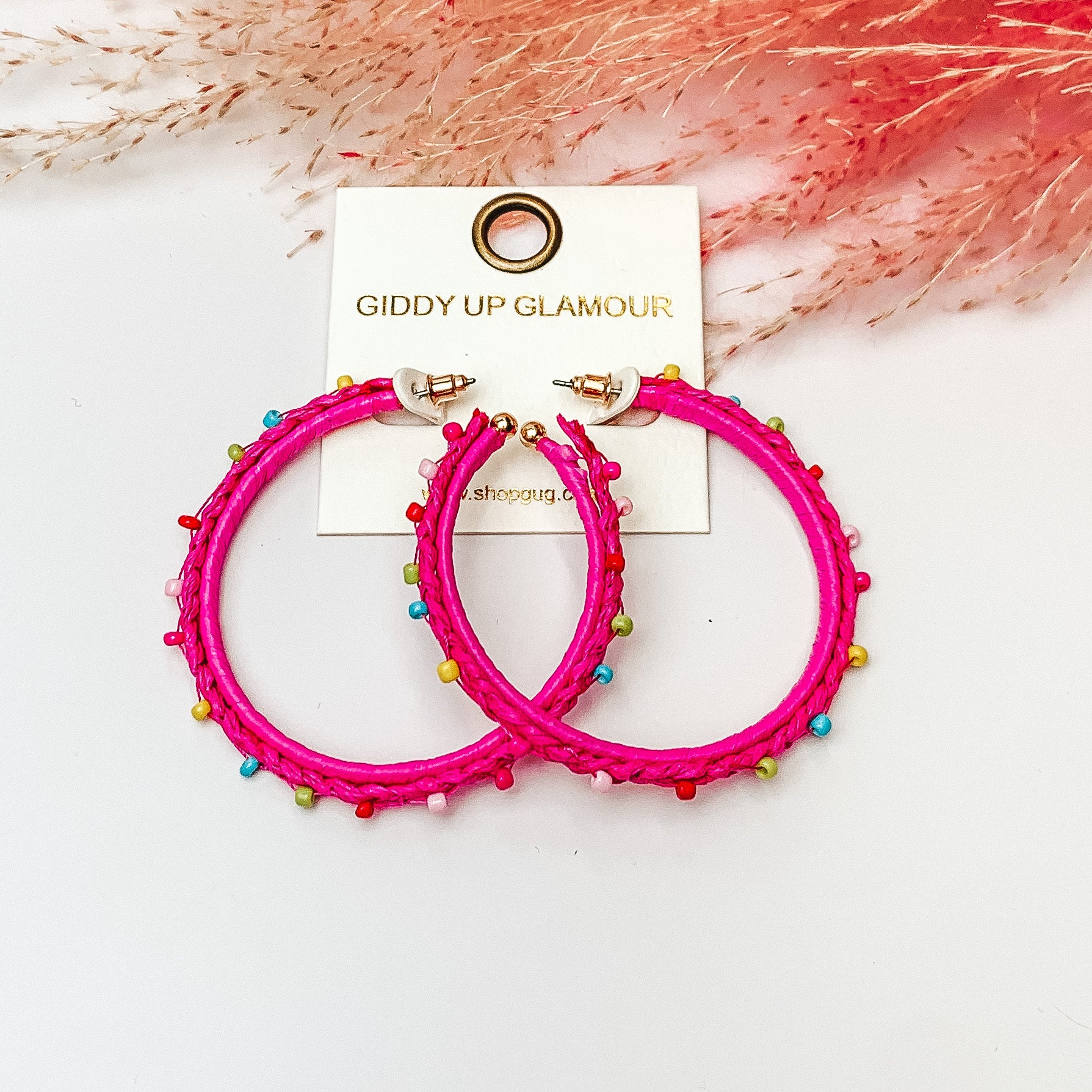 Pictured are raffia braided hoop earrings in fuschia with colorful beads. They are pictured with a pink feather on a white background.
