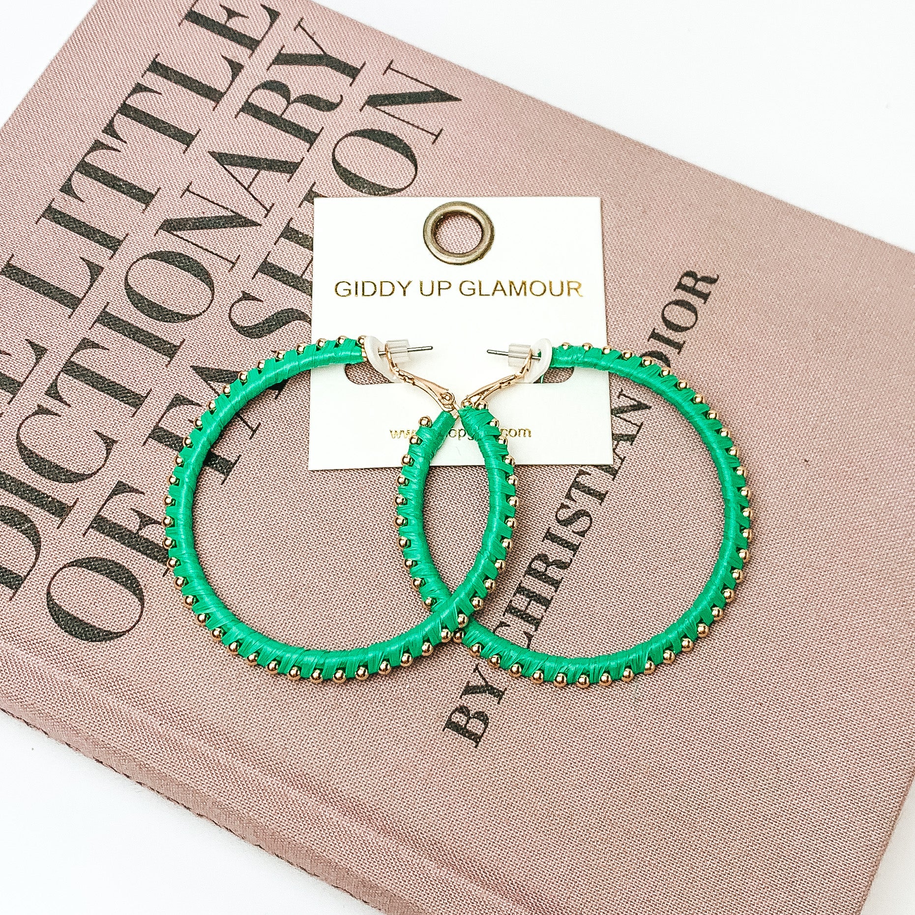 Pictured are circle green hoop earrings with gold beades around it. They are pictured with a pink fashion journal on a white background.