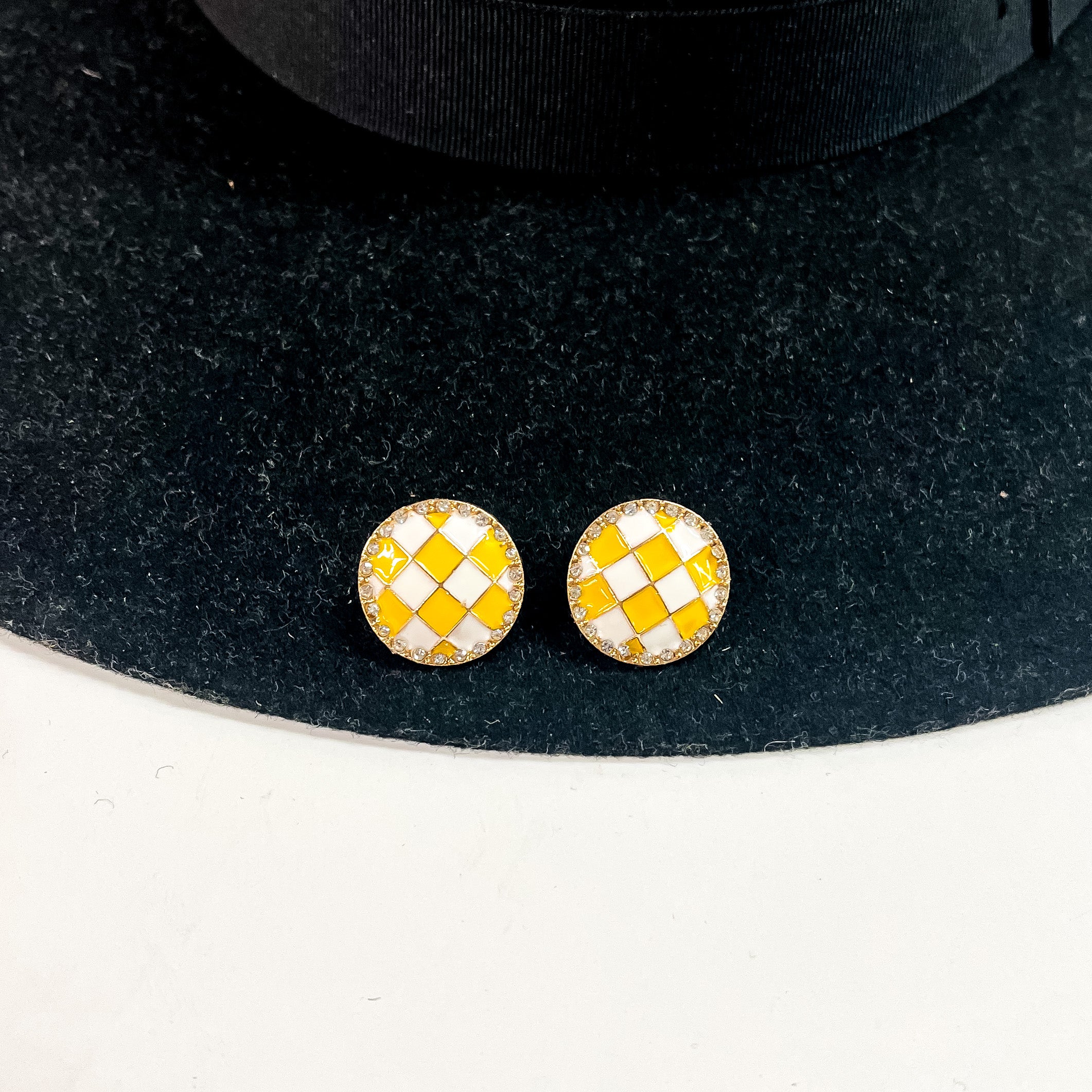 This is a pair of checkered patterned stud earrings in white/yellow in a gold  setting with clear crystals all around. This pair of earrings is laying on a  black felt hat brim and on a white background.