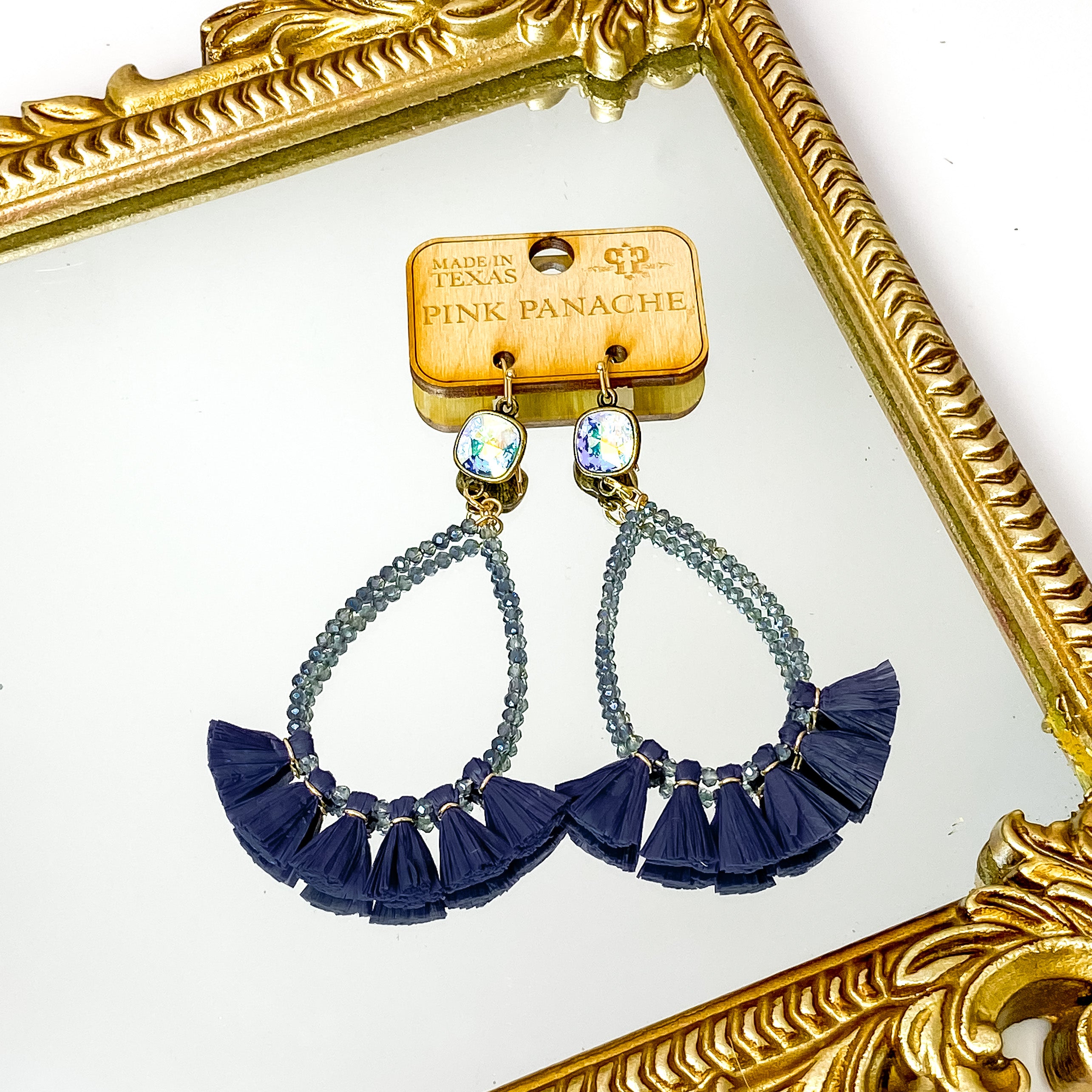 Ab cushion cut drop earrings with a teardrop pendant. The pendant includes navy crystal beads and a navy tassell along the bottom edge. These earrings are pictured on a wood earrings holder on a gold mirror on a white background. 