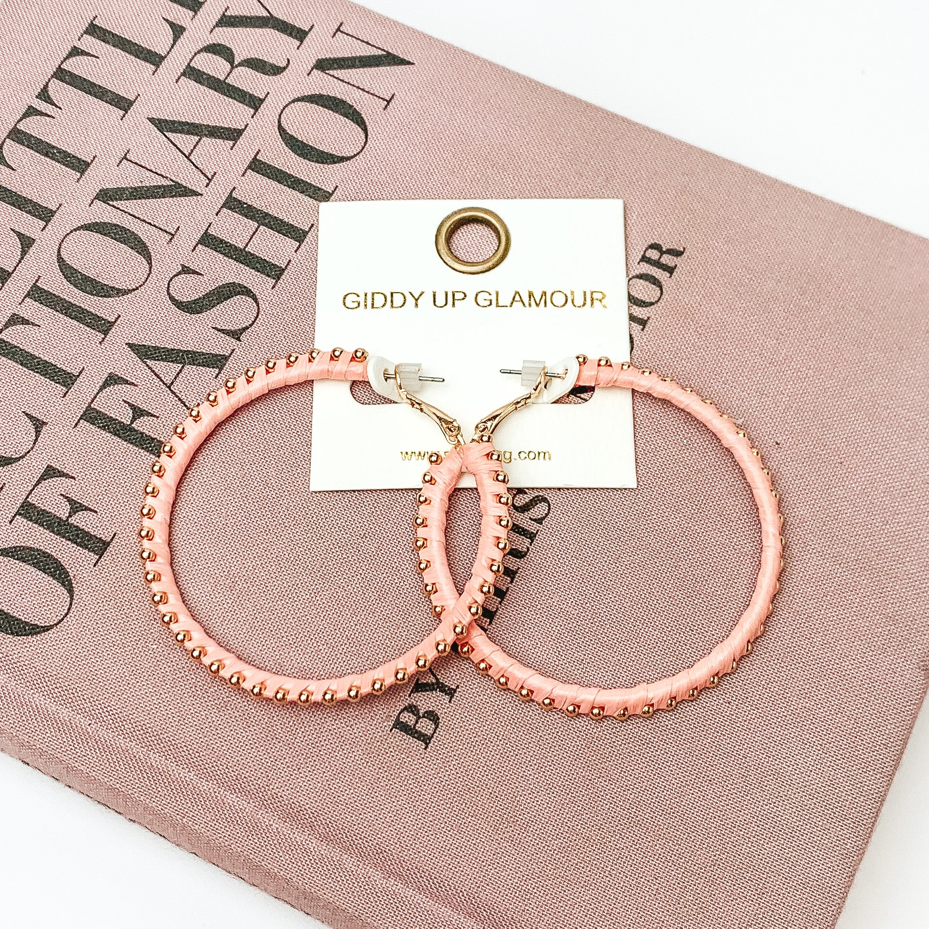  Pictured are circle light pink hoop earrings with gold beads around it. They are pictured with a pink fashion journal on a white background.