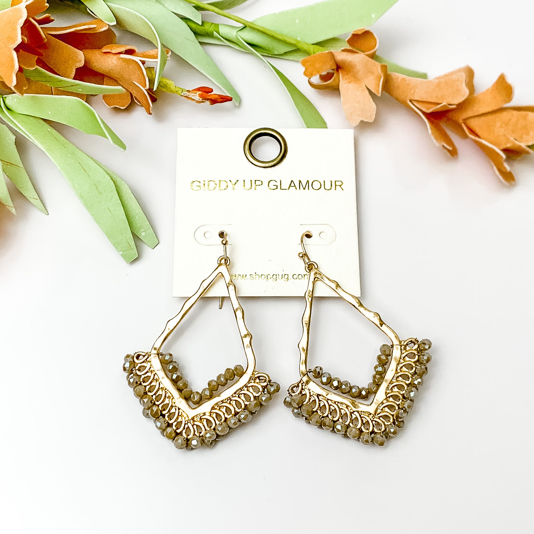 Beige crystal beads bordering open drop gold tone earrings. Pictured on a white background with flowers at the top.