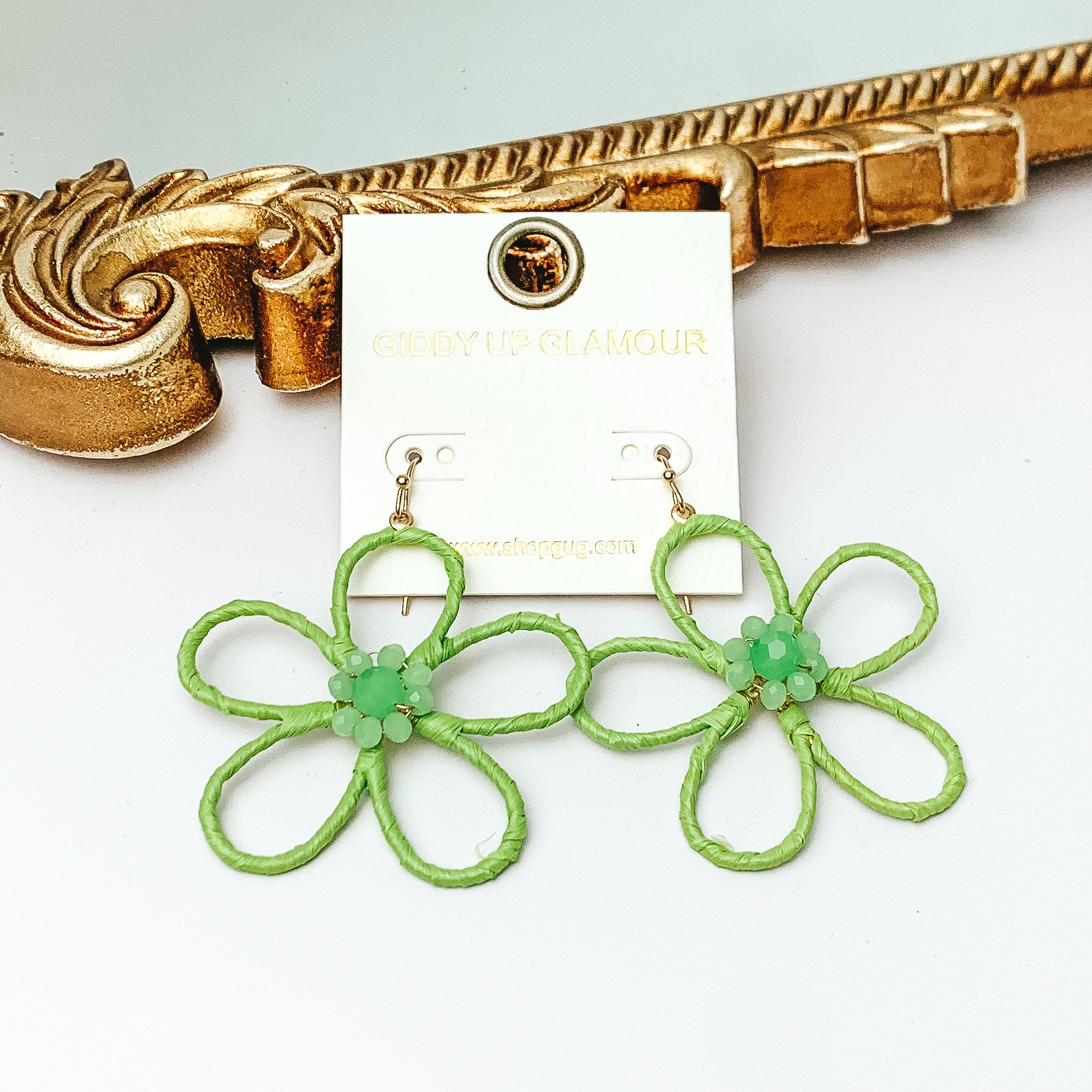 Pictured are gold fish hook earrings with a flower outline pendant. This pendant is pale green and has center pale green crystals. These earrings are pictured in front of a gold mirror on a white background. 