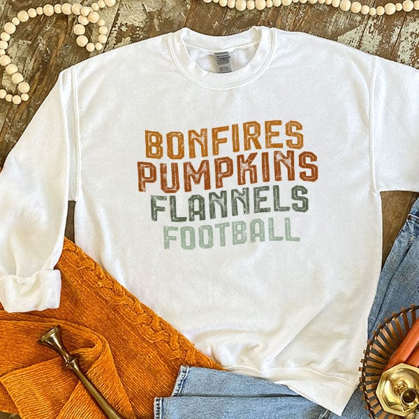 This white sweatshirt includes a crew neckline, long sleeves, and cute hand drawn design of the following words stacked on top of each other, "Bonfires Pumpkins Flannels, Football" in fun, Fall colors. This is shown here as a flat lay with rolled sleeves and light wash denim jeans.