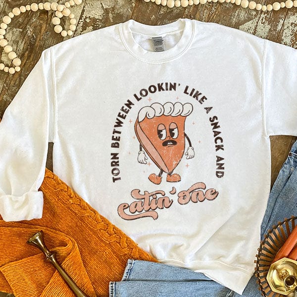 This white sweatshirt includes a crew neckline, long sleeves, and a cute hand drawn design of a slice of pie with the words "Torn Between Lookin' Like A Snack And Eatin' One" around it. It is shown styled as a flat lay in this photo with the sleeves rolled and paired with light wash denim jeans.  