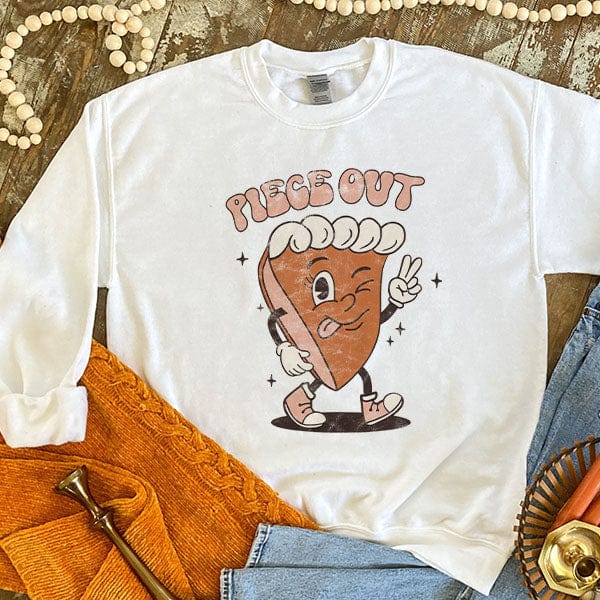 This white sweatshirt includes a crew neckline, long sleeves, and cute hand drawn design of a piece of pie with hands, legs, and feet. The pie's left hand is holding up the peace sign with the words "Piece Out" above it. This is shown as a flat lay with light wash denim jeans and the sleeves are shown rolled. 