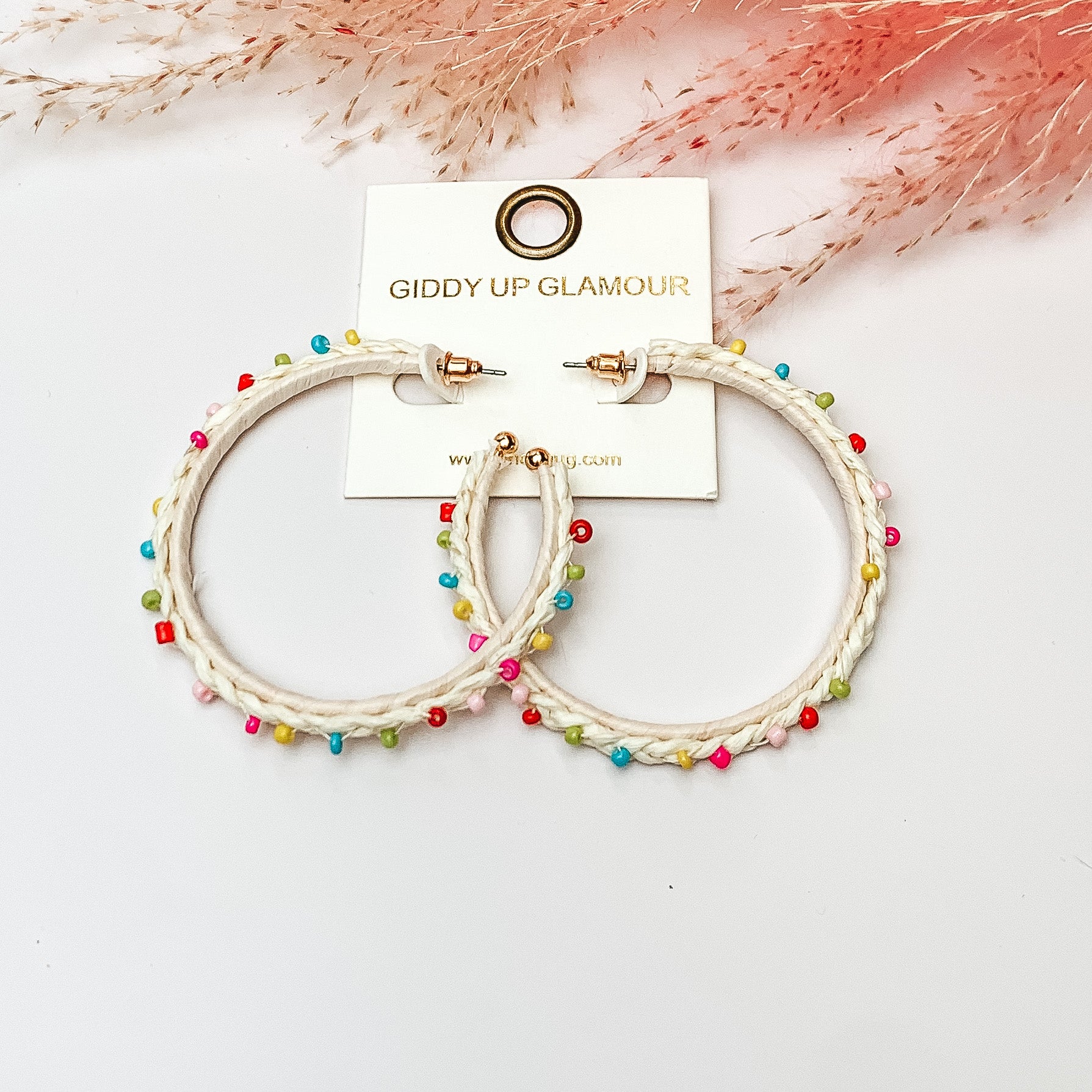 Pictured are raffia braided hoop earrings in ivory with colorful beads. They are pictured with a pink feather on a white background.