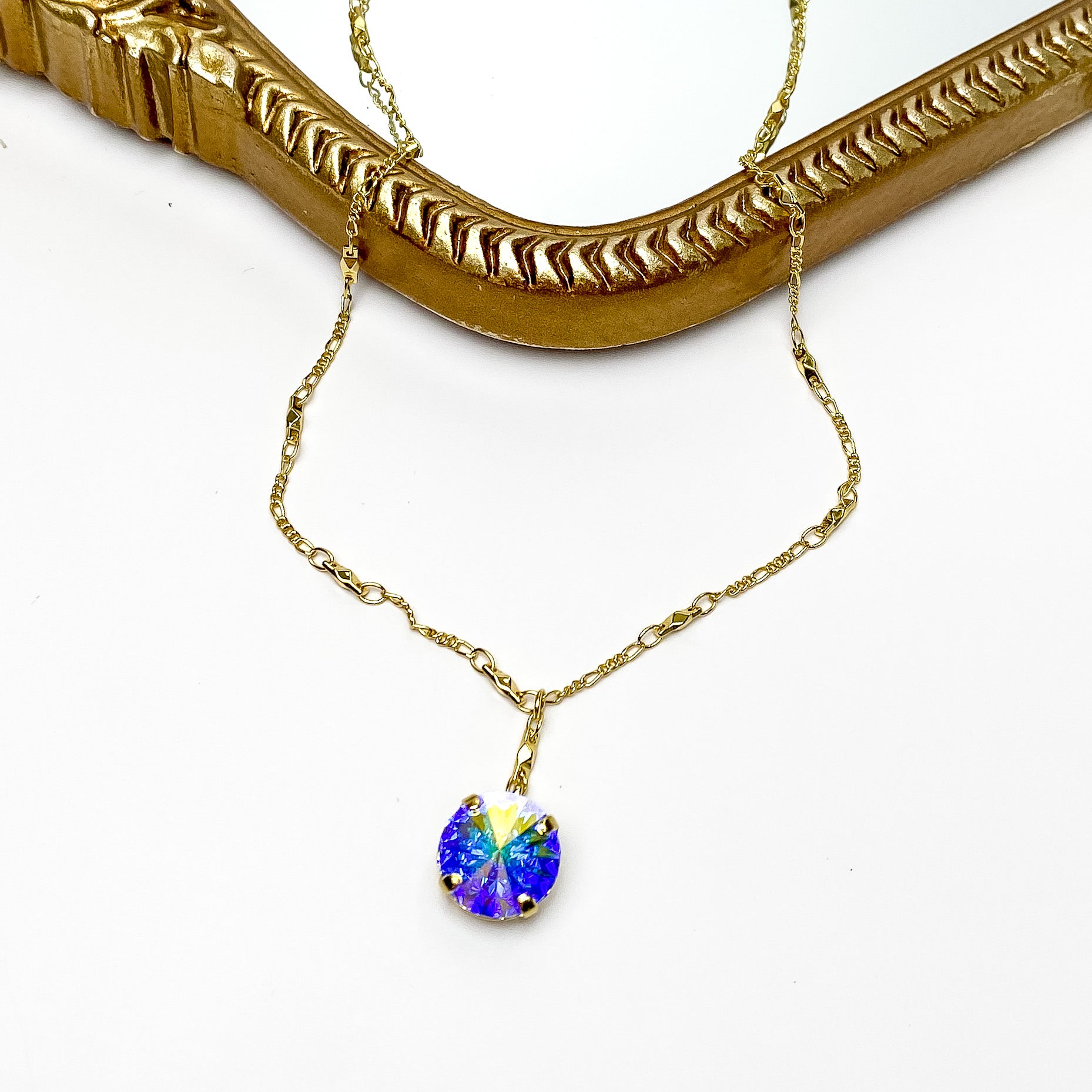 Pictured is a gold necklace with a ab crystal pendant. This necklace is pictured laying partially on a gold mirror on a white background. 