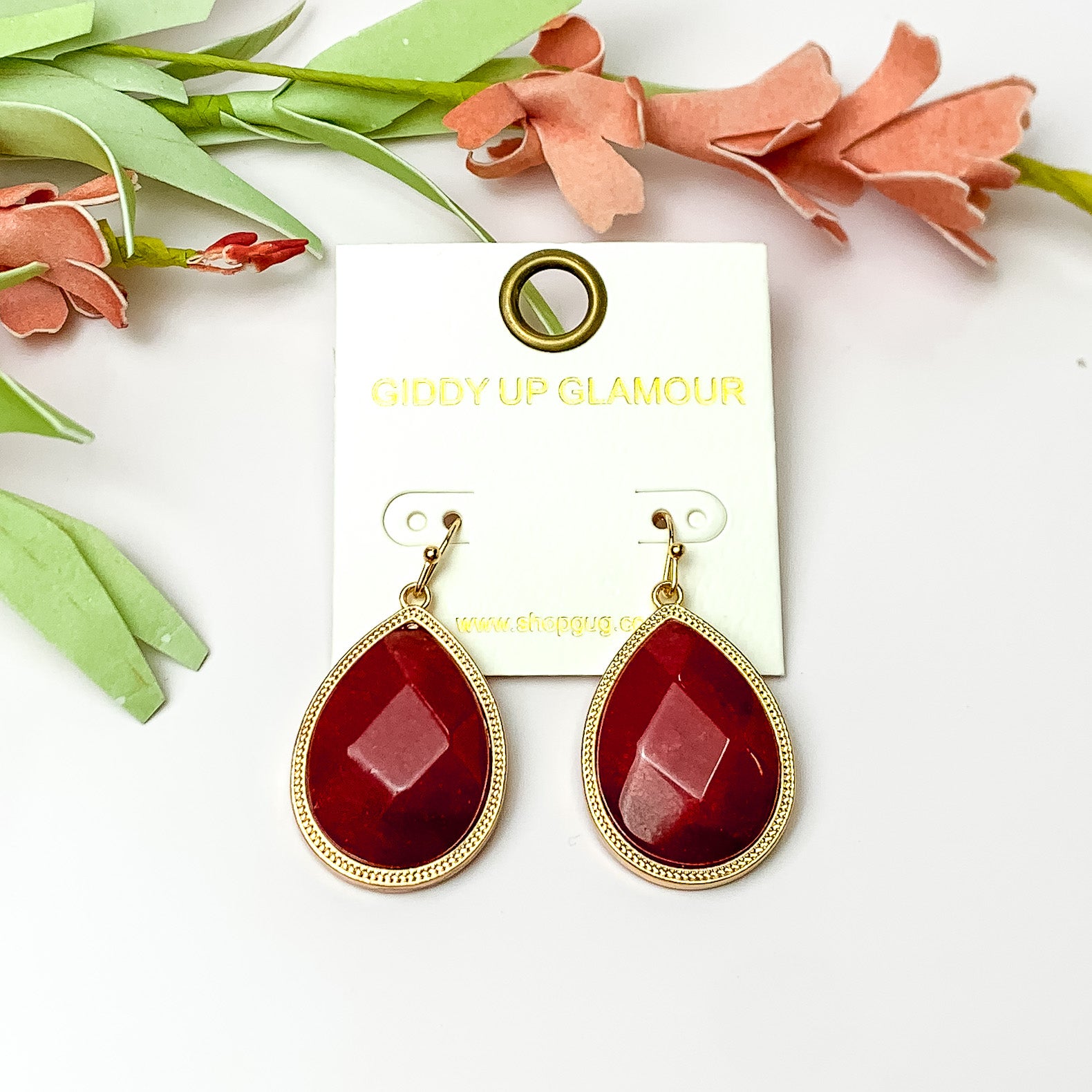 Maroon drop earrings with gold tone outline. Pictured on a white background with flowers at the top.