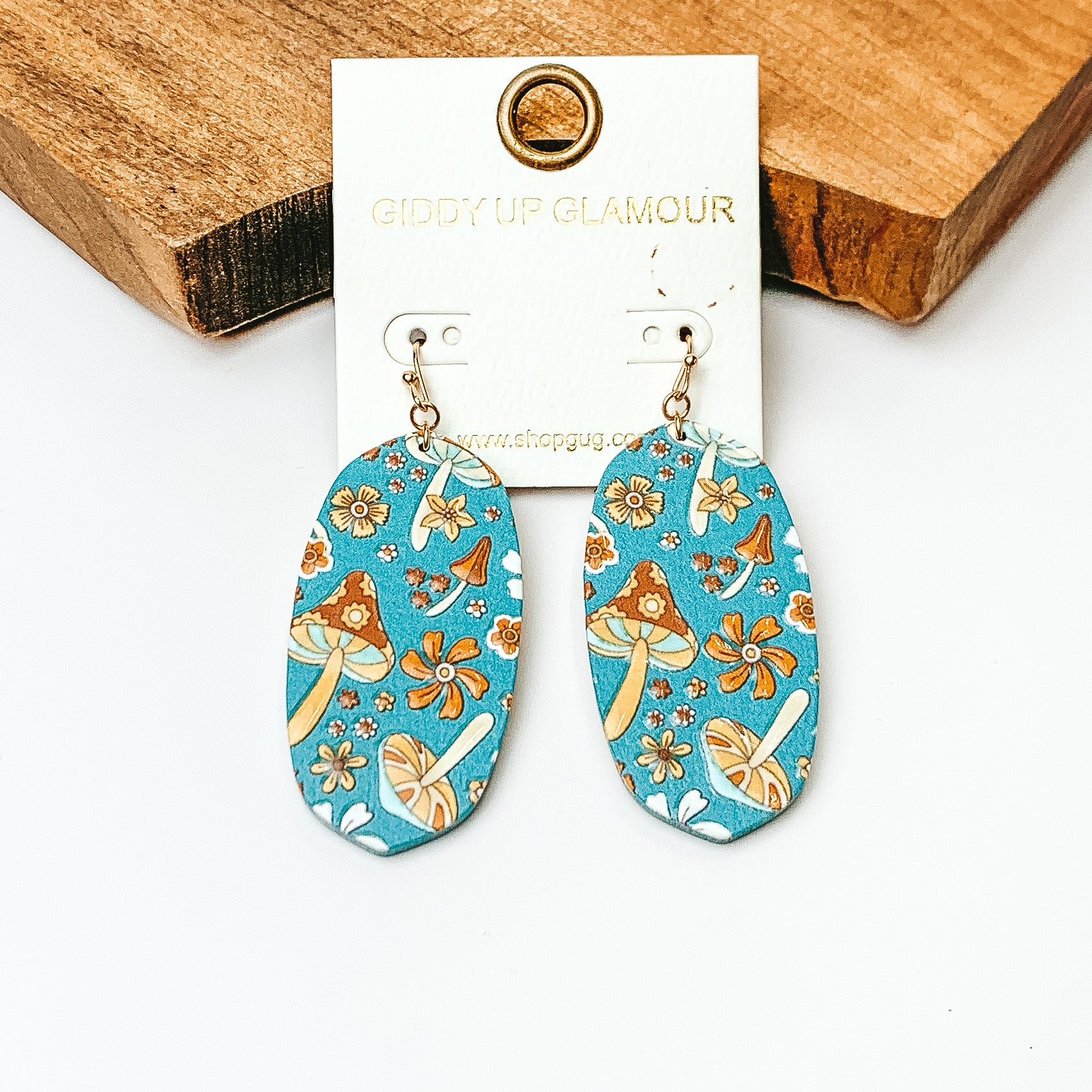 Pictured are oval drop earrings on gold fish hooks. These earrings have a blue background with a mix of flowers and mushrooms print. These earrings are pictured on a white background in front of a wood block. 