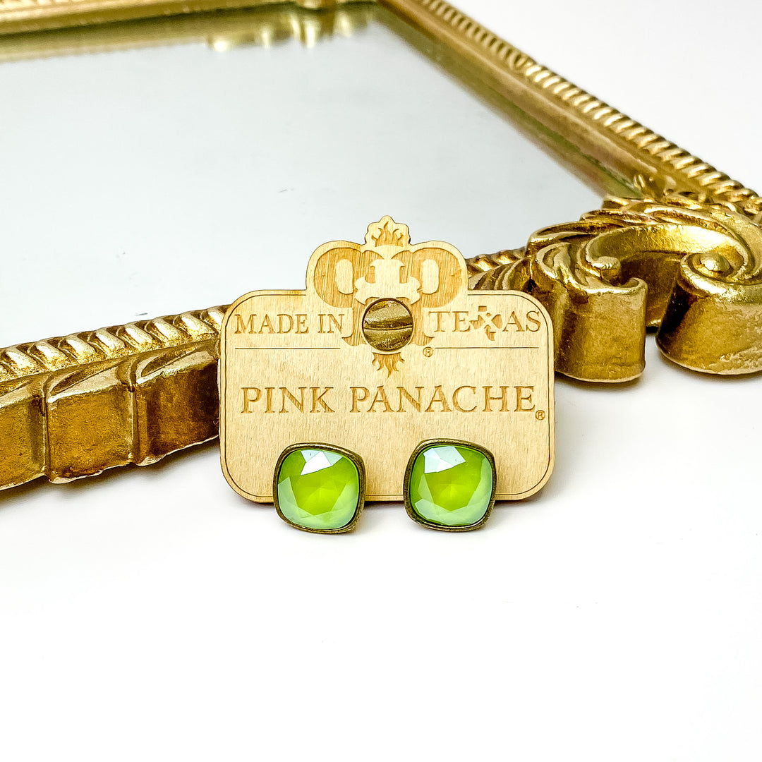 Lime green cushion cut crystal stud earrings with a bronze setting. These earrings are pictured on a Pink Panache wood holder in front of a gold mirror and on a white background. 