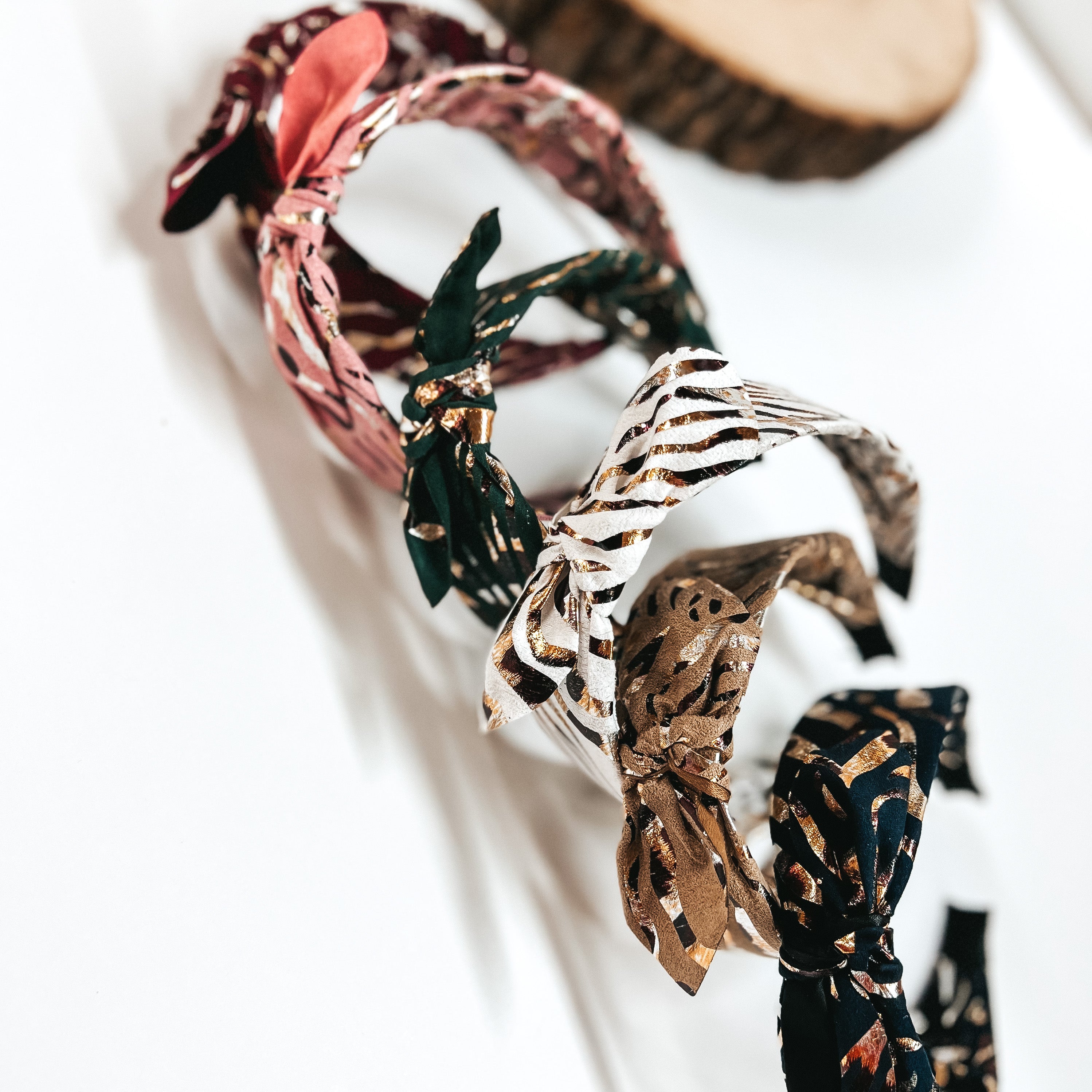 There are six fabric headbands with a bow and gold foil  print in different colors. From top to bottom; burgundy, light pink, dark green, white, tan, and black.