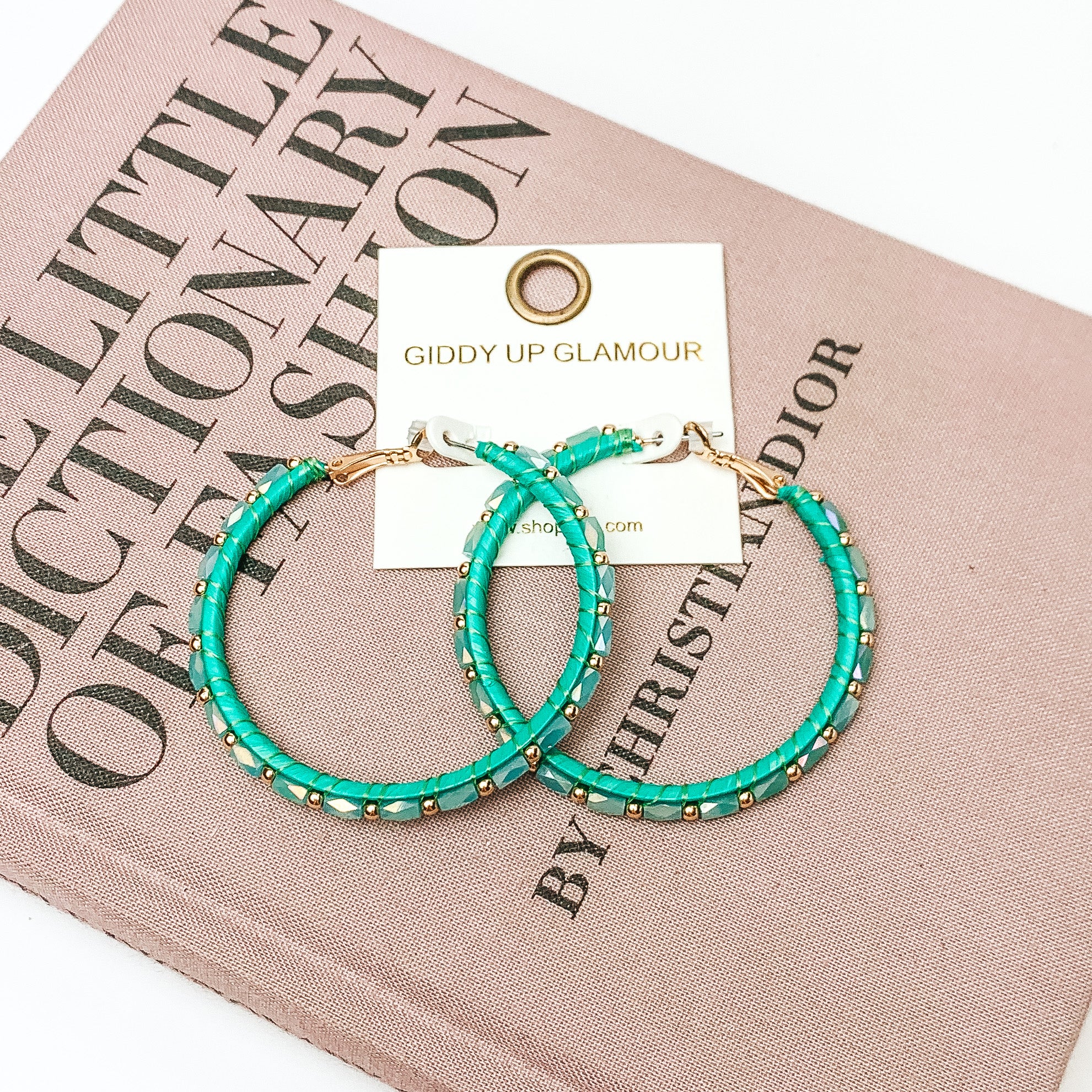Pictured are circle beaded hoop earrings with gold spacers in green. They are pictured with a pink fashion journal on a white background
