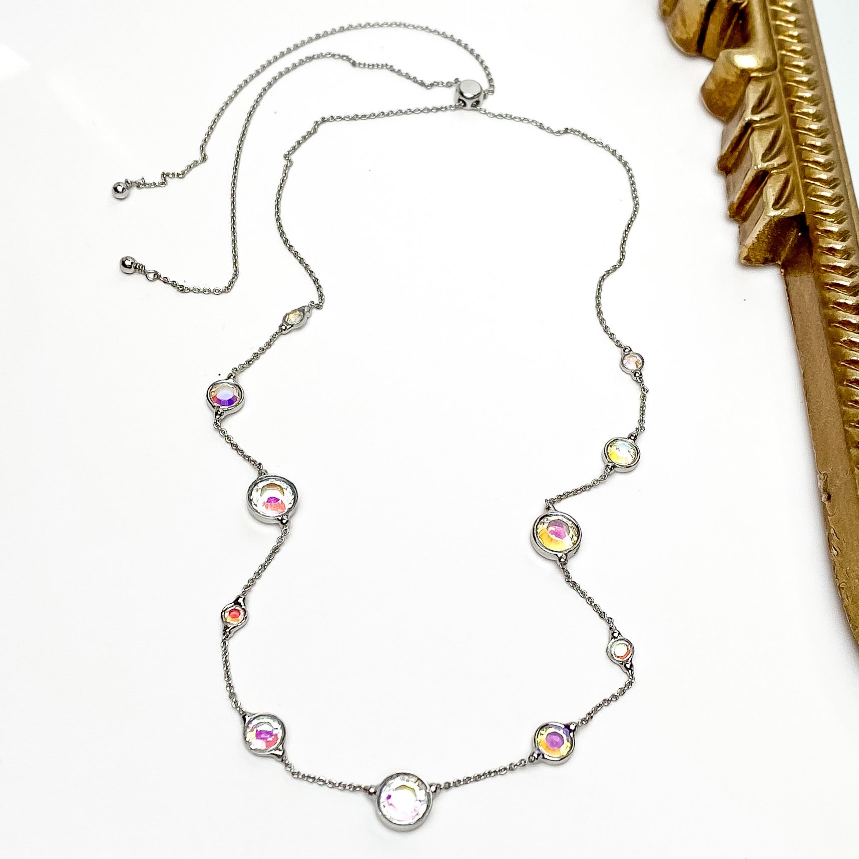 Pictured is a thin, silver chain with spaced out circle ab crystals. These crystals also come in different sizes. This necklace is pictured on a white background with a gold mirror to the right of the necklace.    