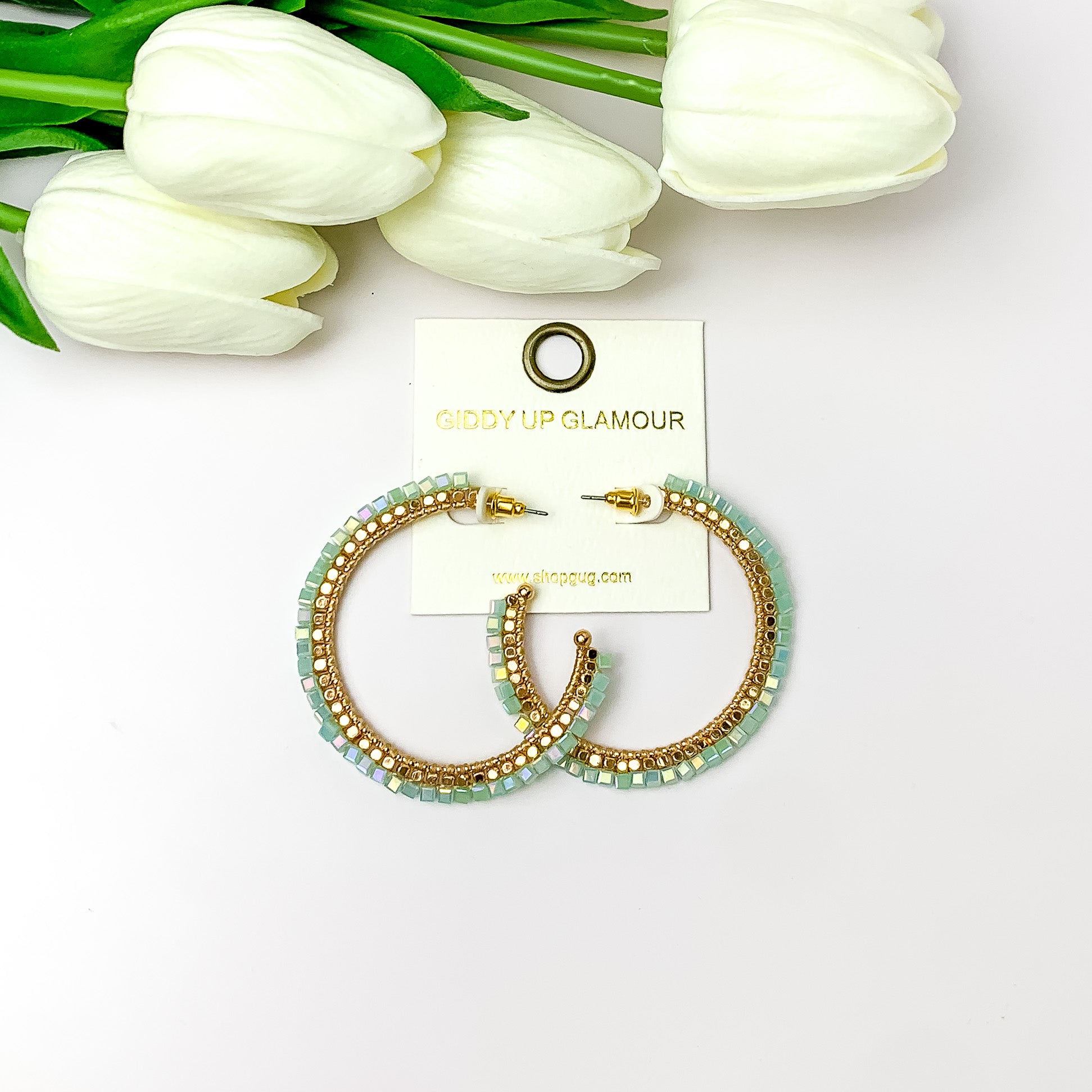 Pictured are circle gold toned hoop earrings with gold beads around it and mint crystal outline. They are pictured with white flowers on a white background.