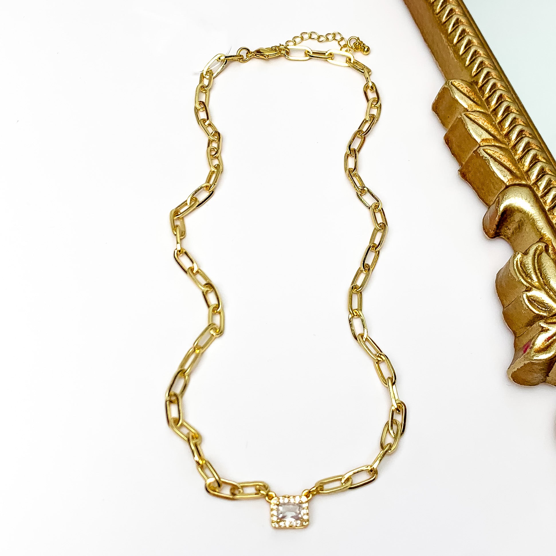 Pictured is a gold chain necklace with a rectangle pendant with clear crystals. This necklace is pictured on a white background with a gold mirror in the top right corner.  