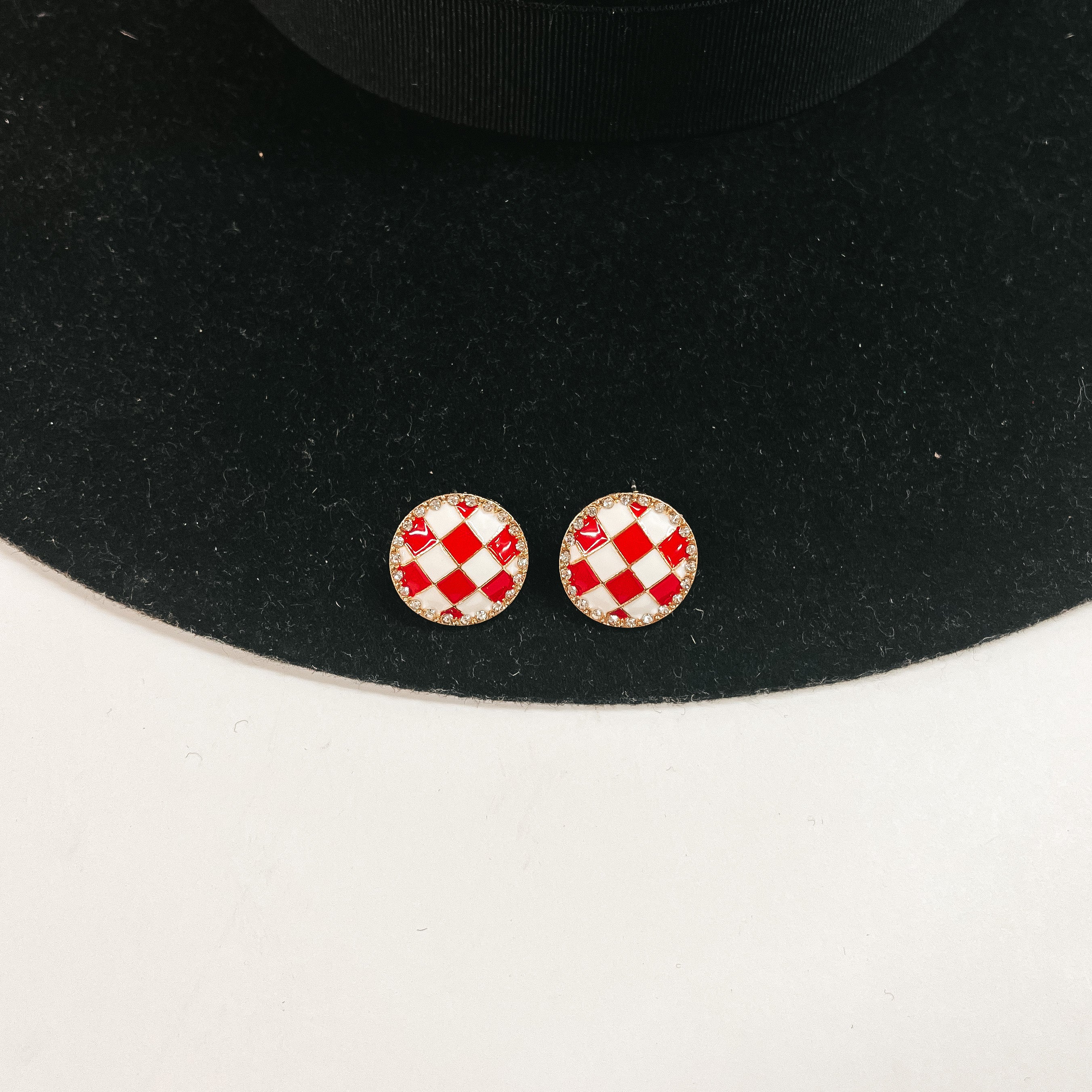 This is a pair of checkered patterned stud earrings in white/red in a gold  setting with clear crystals all around. This pair of earrings is laying on a  black felt hat brim and on a white background.