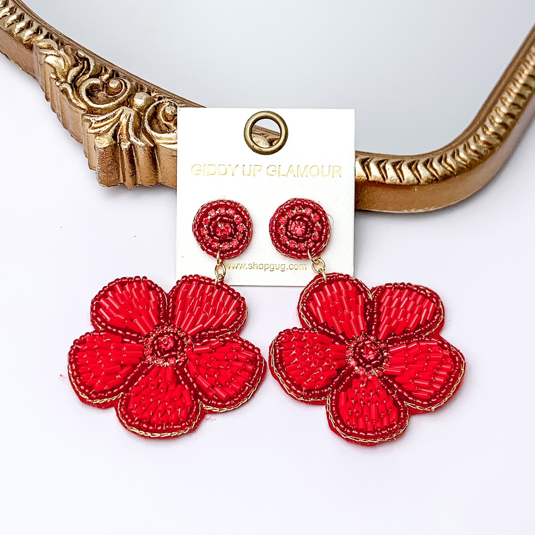 There is hanging beaded flower pendant from the gold stud earrings. The flower pendants are red with gold outline and detailing. These earrings are pictured in front of a gold mirror on a white background.