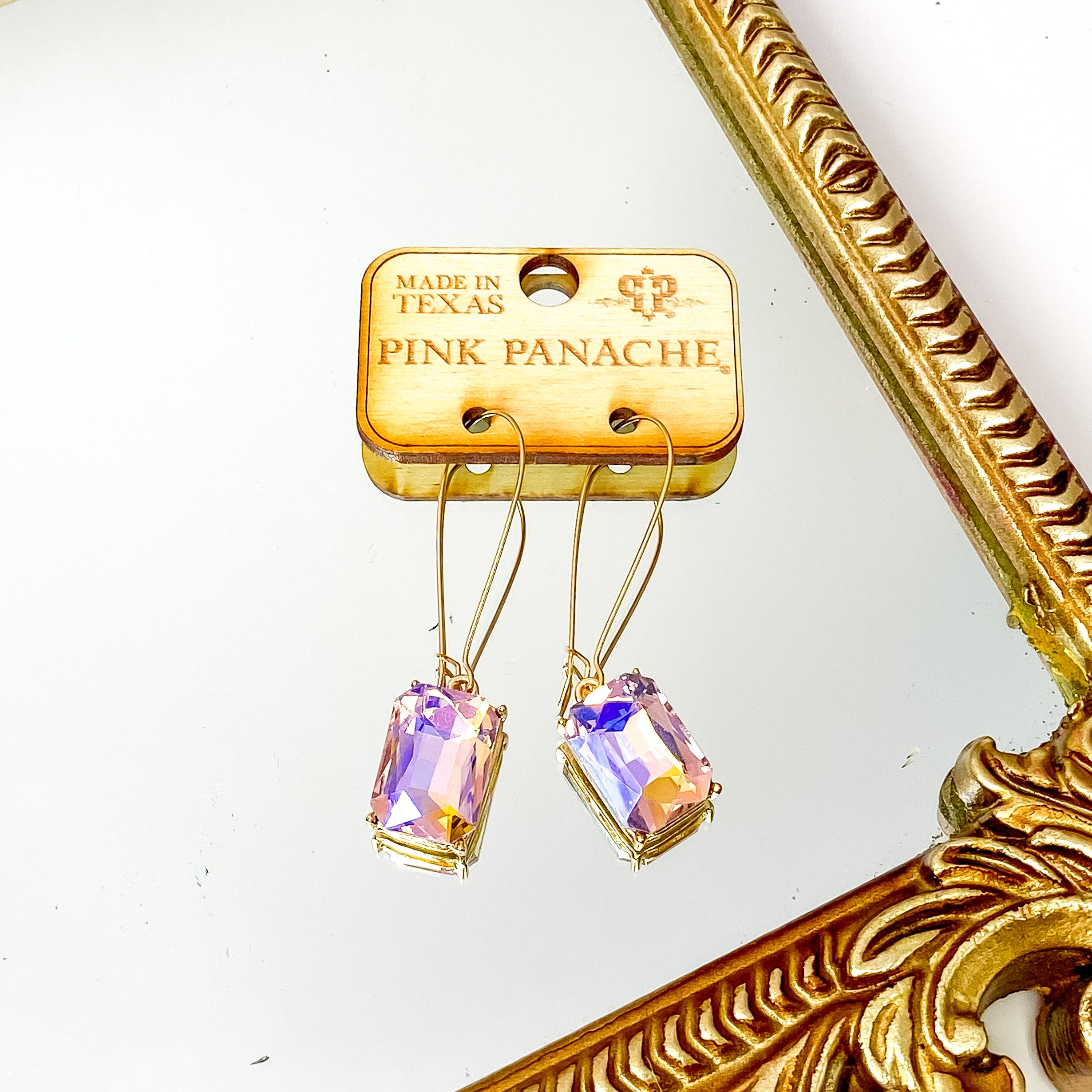 Gold kidney wire drop earrings with a rose ab rectangle crystal charm. These earrings are pictured on a wood earrings holder in front of a gold mirror on a white background. 