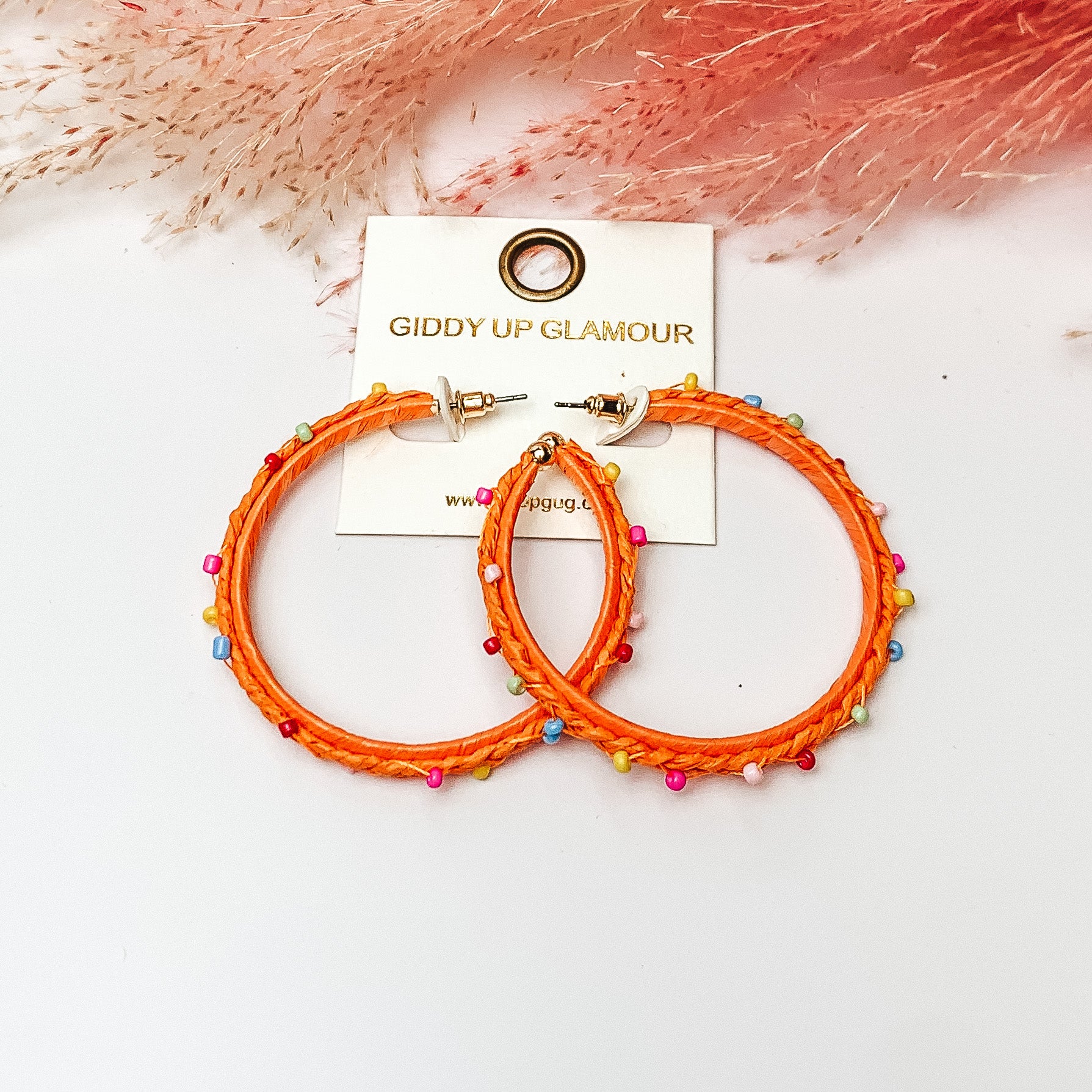 Pictured are raffia braided hoop earrings in orange with colorful beads. They are pictured with a pink feather on a white background.