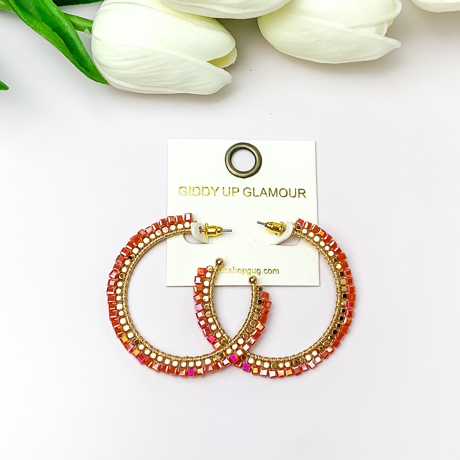  Pictured are circle gold toned hoop earrings with gold beads around it and a red orange crystal outline. They are pictured with white flowers on a white background.