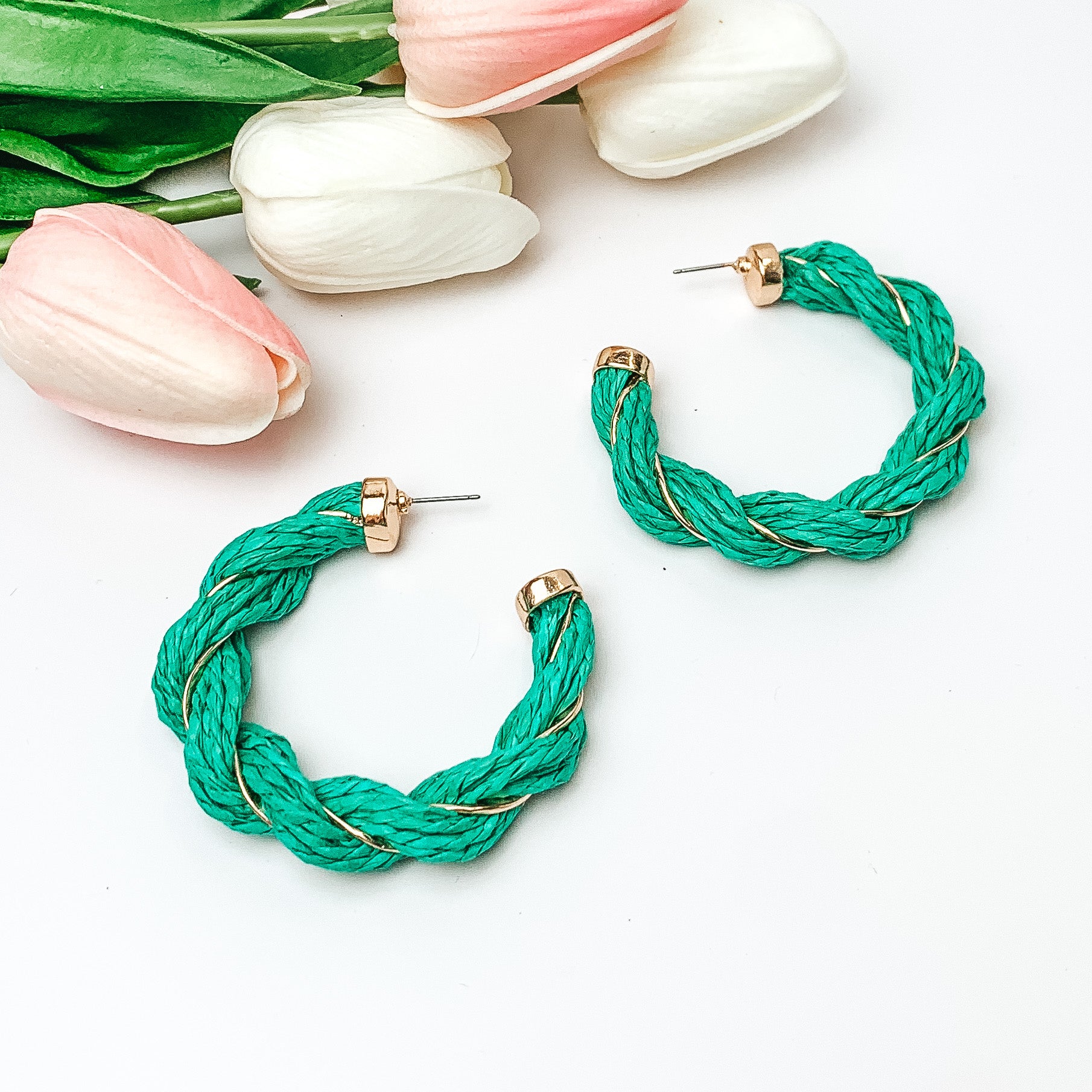 Pictured are teal raffia twisted hoop earrings with gold detailing.  They are pictured with pink and white tulips on a white background.