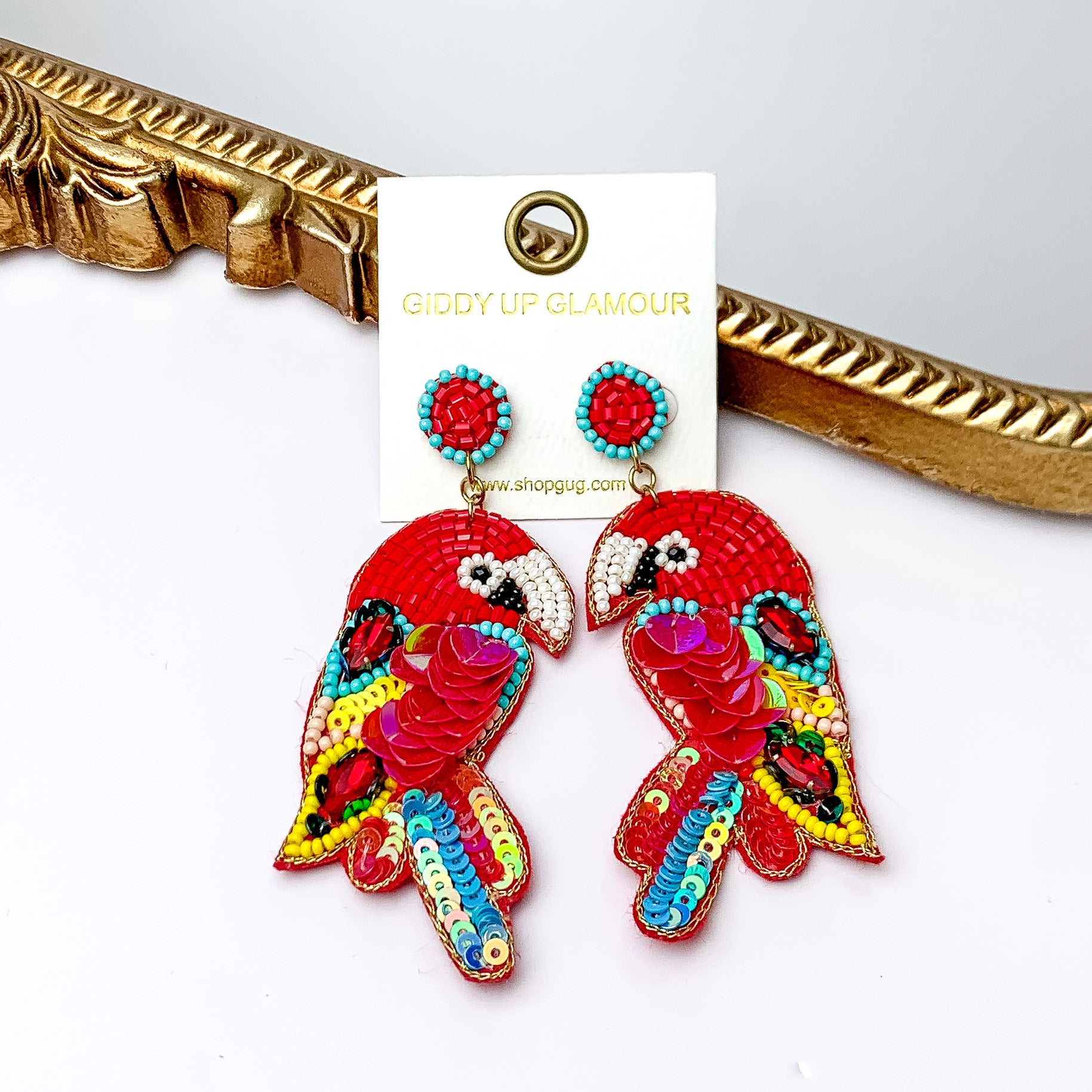 These parrot earrings are red with a little bit of blue, yellow, and black beads. There is also some red  and green seqence across the earrings. These earrings are pictured in front of a gold mirror on a white background.