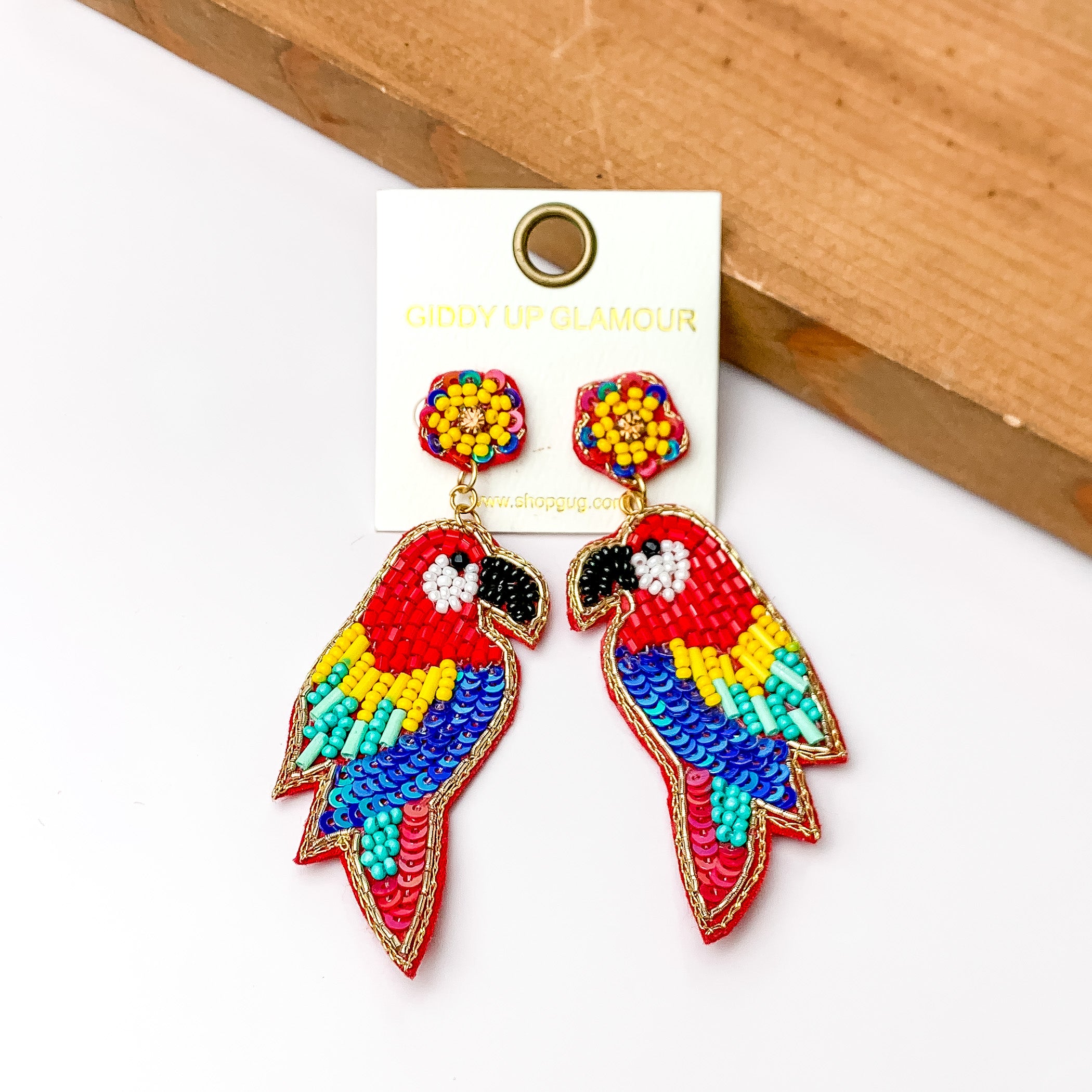These parrot earrings are red with beads that are yellow, turquoise, and black. They do have seqence that are red and royal blue. These earrings are pictured in front of a piece of wood with a white background.