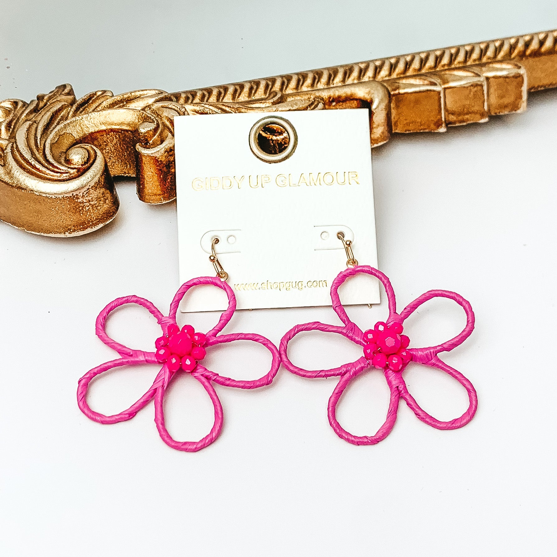 Pictured are gold fish hook earrings with a flower outline pendant. This pendant is fuchsia pink and has center fuchsia pink crystals. These earrings are pictured in front of a gold mirror on a white background. 