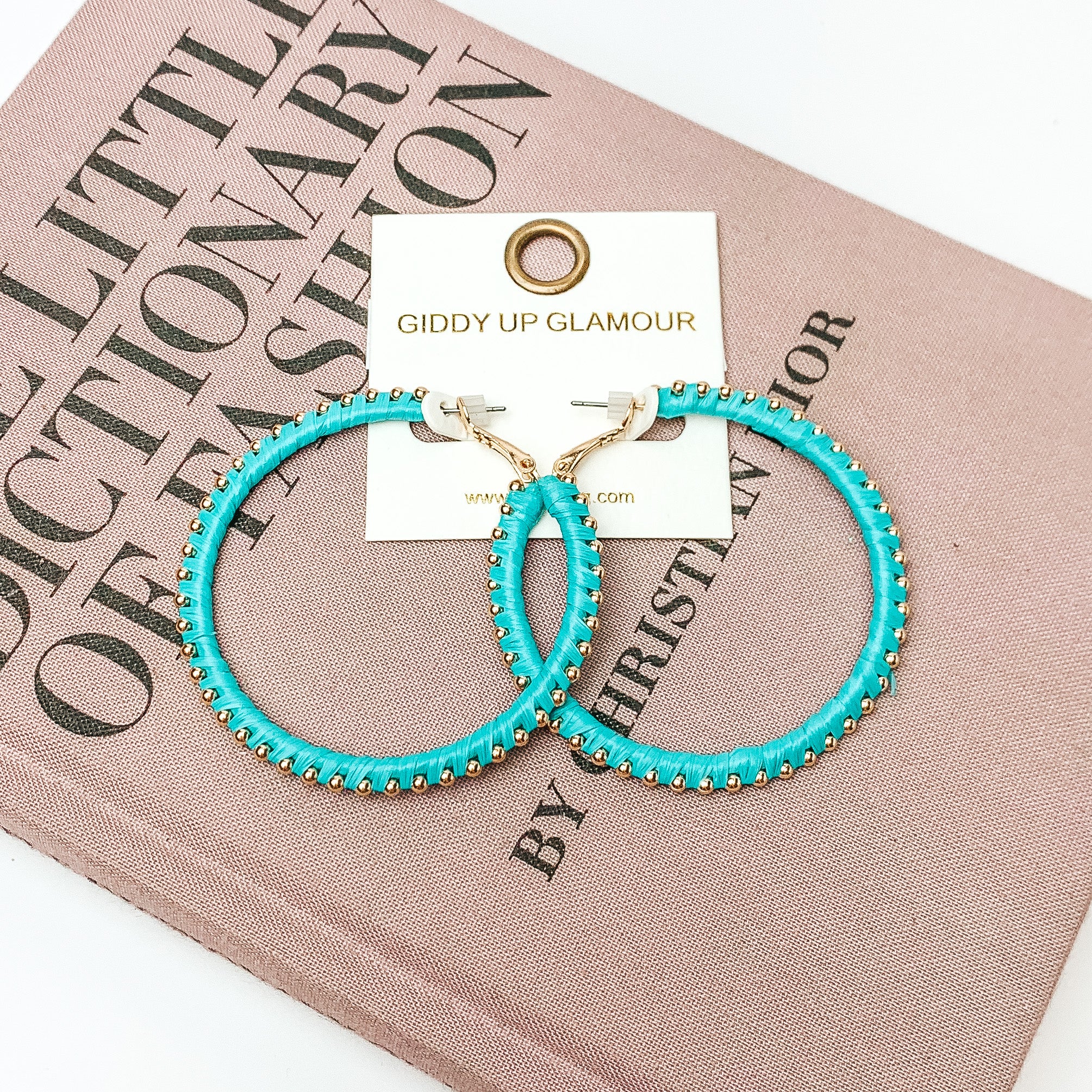 Pictured are circle turquoise hoop earrings with gold beads around it. They are pictured with a pink fashion journal on a white background.