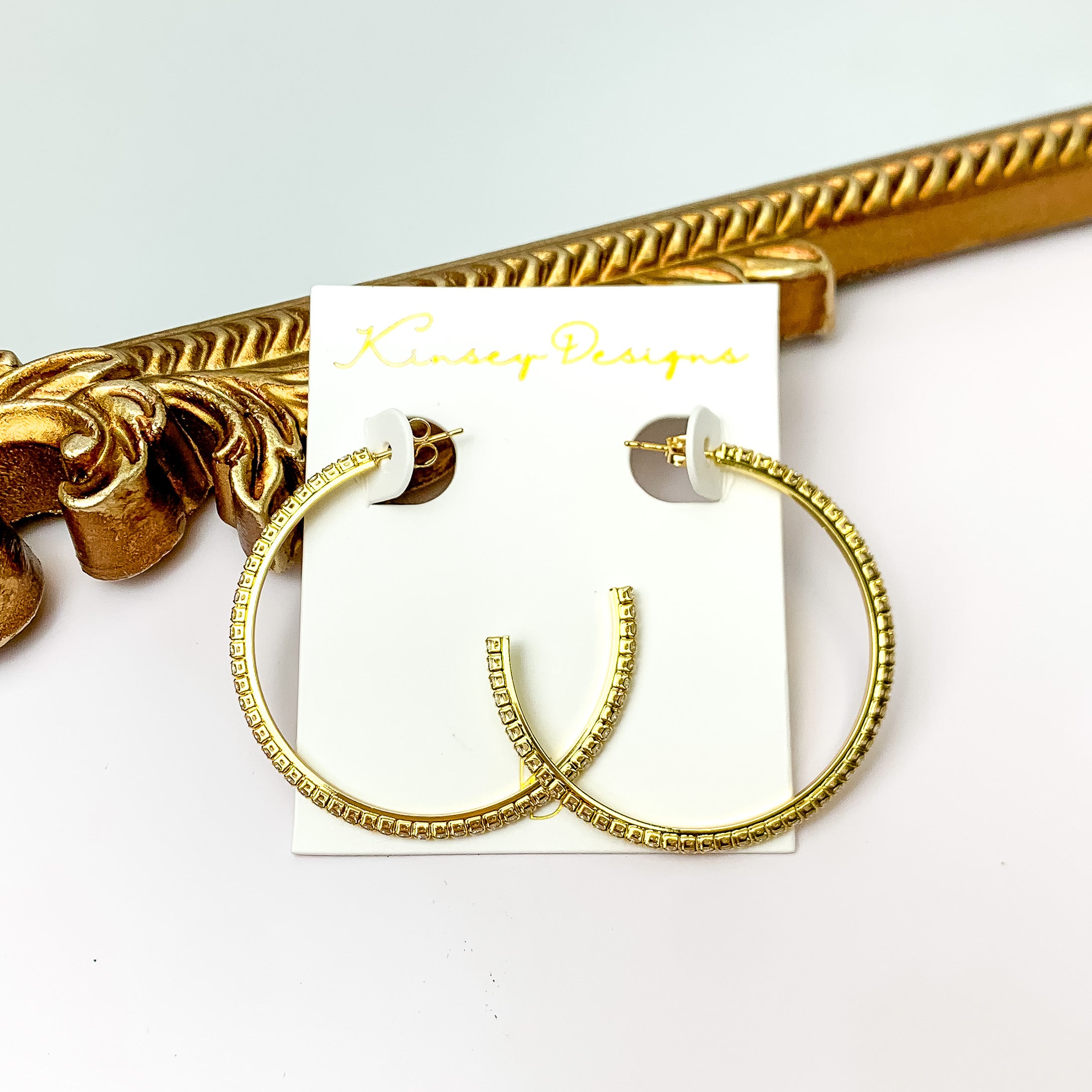 A pair of gold hoop earrings outlined with clear crystals. These earrings are pictured in front of a gold mirror on a white background.  