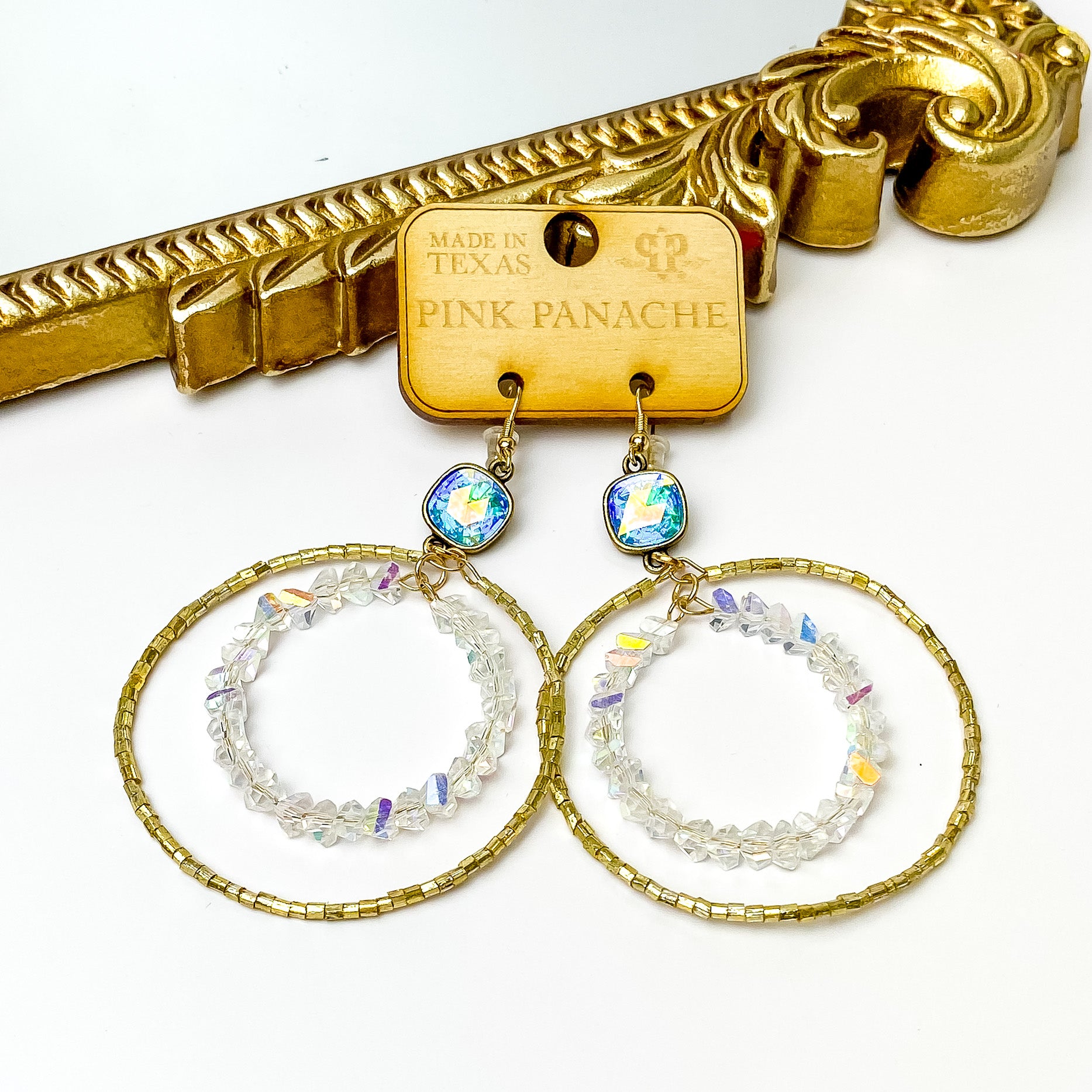 Ab cushion cut crystal drop earrings with a double circle pendant. One circle is gold beaded and the smaller one has clear ab crystal beads. These earrings are pictured on a wood earrings holder in front of a gold mirror on a white background. 