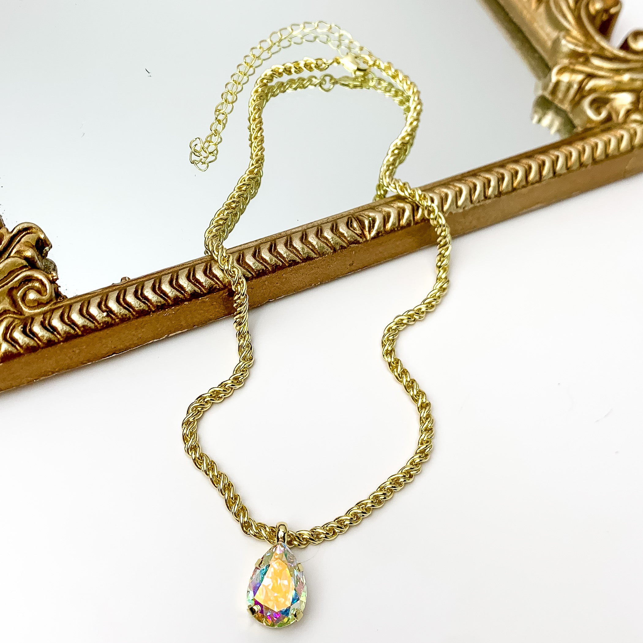 Pictured is a gold chain necklace with ab crystal teardrop charm. This necklace is pictured partially laying on a gold mirror on a white background.    