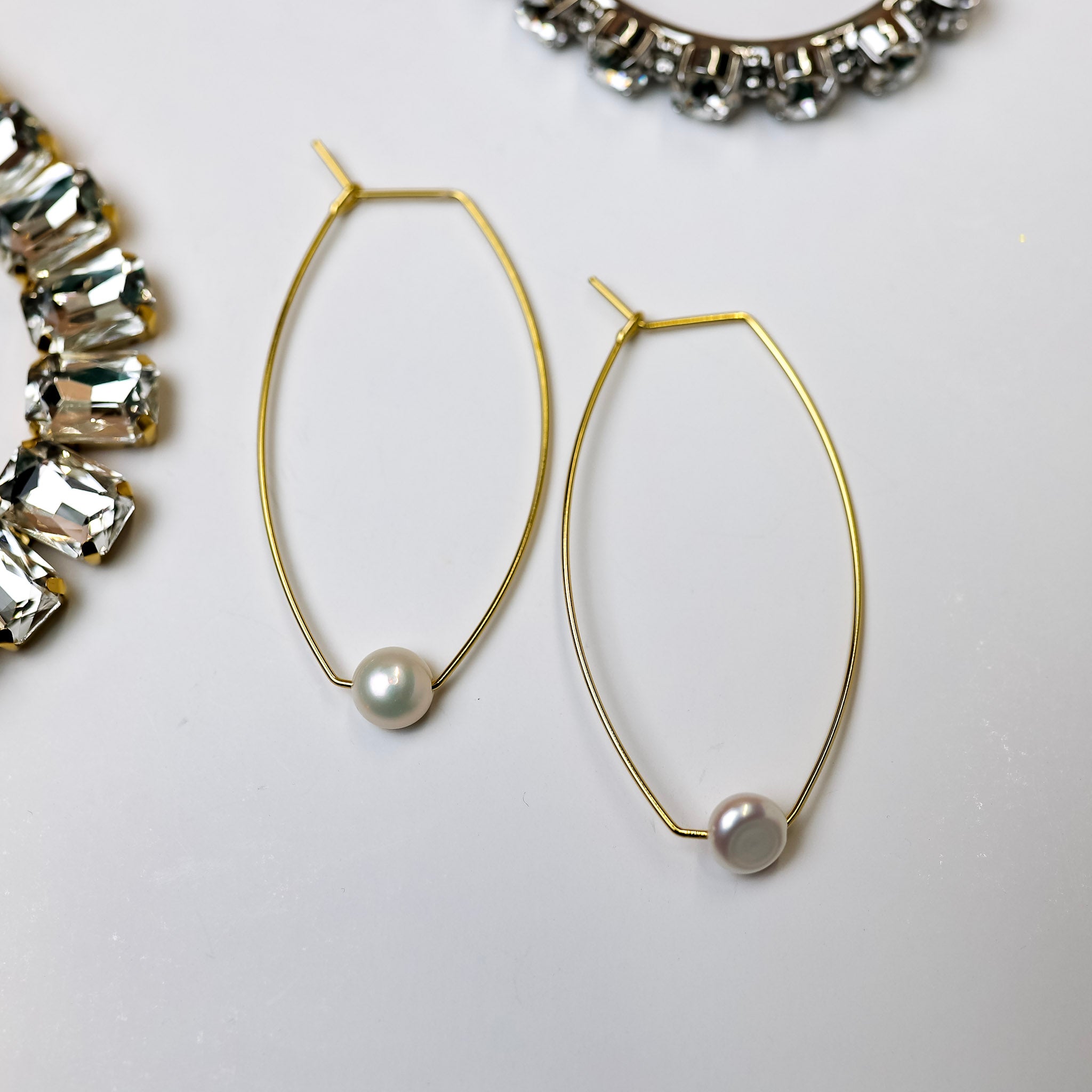 A pair of gold-tone cat eye hoop earrings with a pearl pendant at the bottom of each. Pictured on a white background with crystal necklaces.