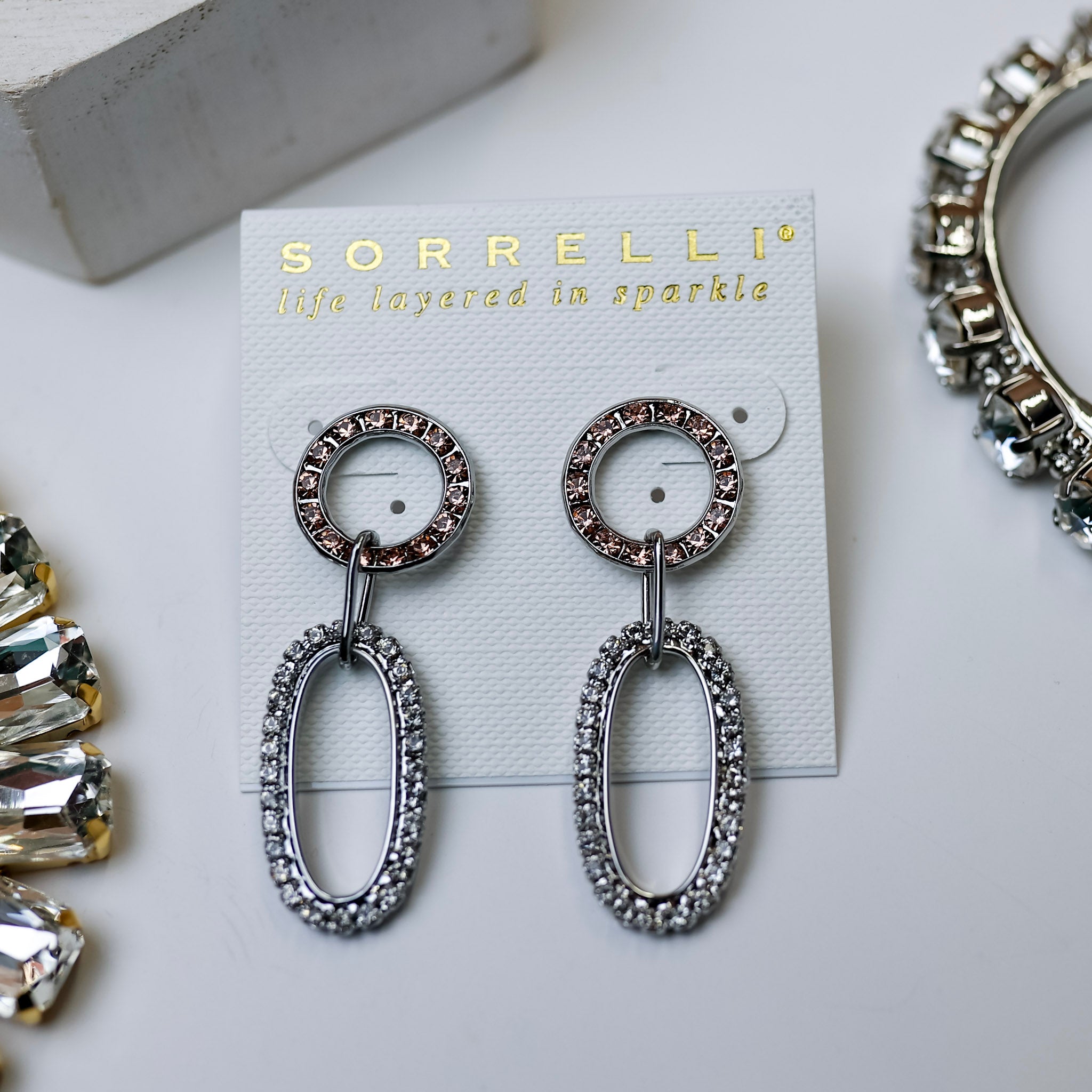 A pair of dangle earrings that consist of a circle outline with blush crystals and an oval outline with clear crystals. These earrings are pictured on a white background with crystal necklaces.