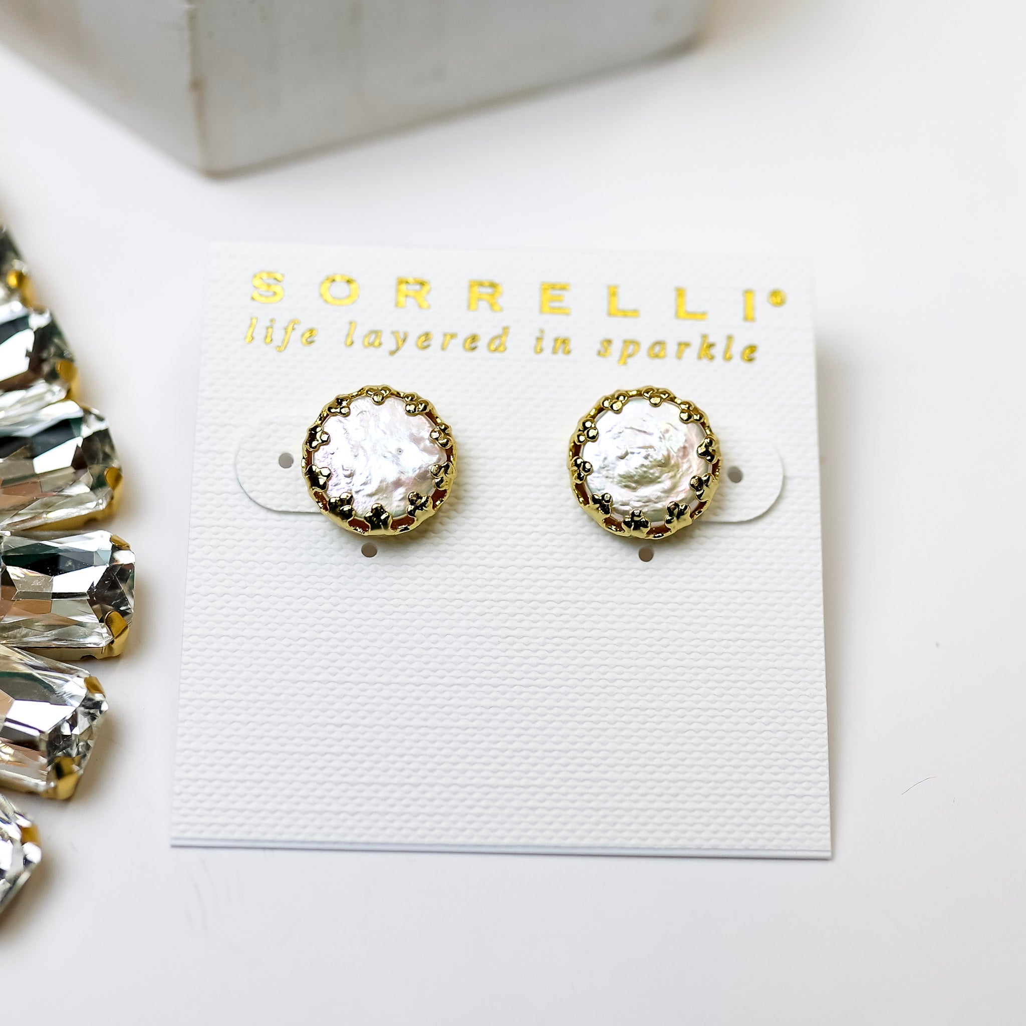 A pair of gold tone circle stud earrings with an ivory pearl center. Pictured on a white background with crystal necklaces.