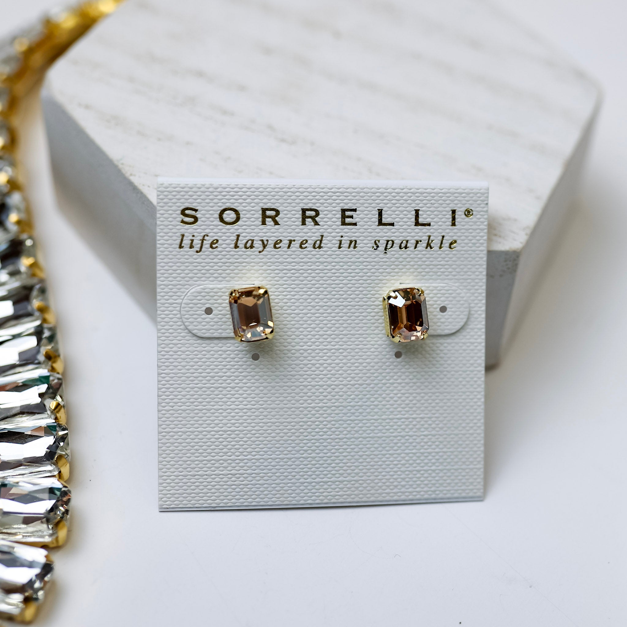 A pair of small emerald cut crystal stud earrings in dark champagne pictured on a white background with crystal necklaces.
