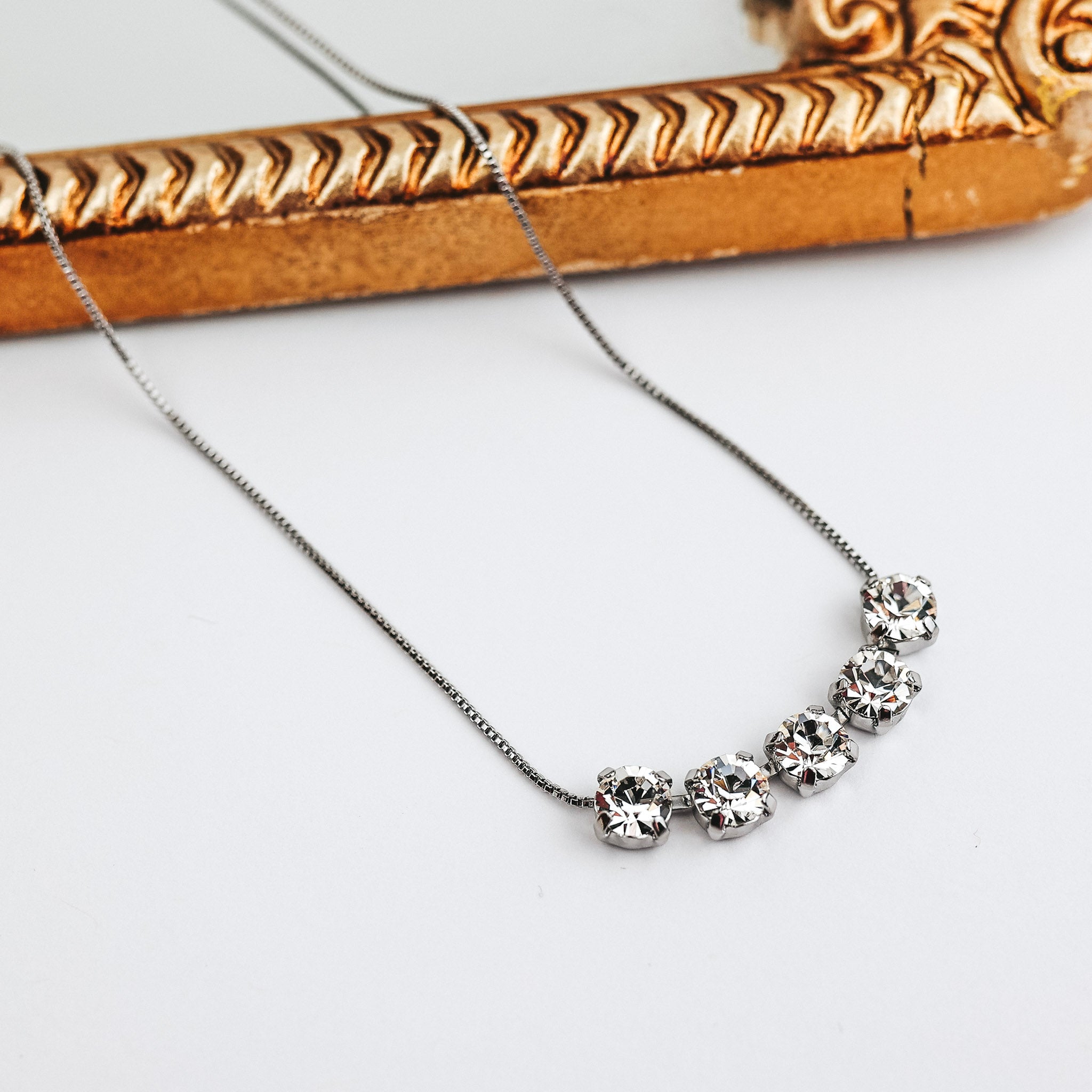 A silver tone necklace with five clear crystals at the front. This necklace is pictured on a white background with a mirror.