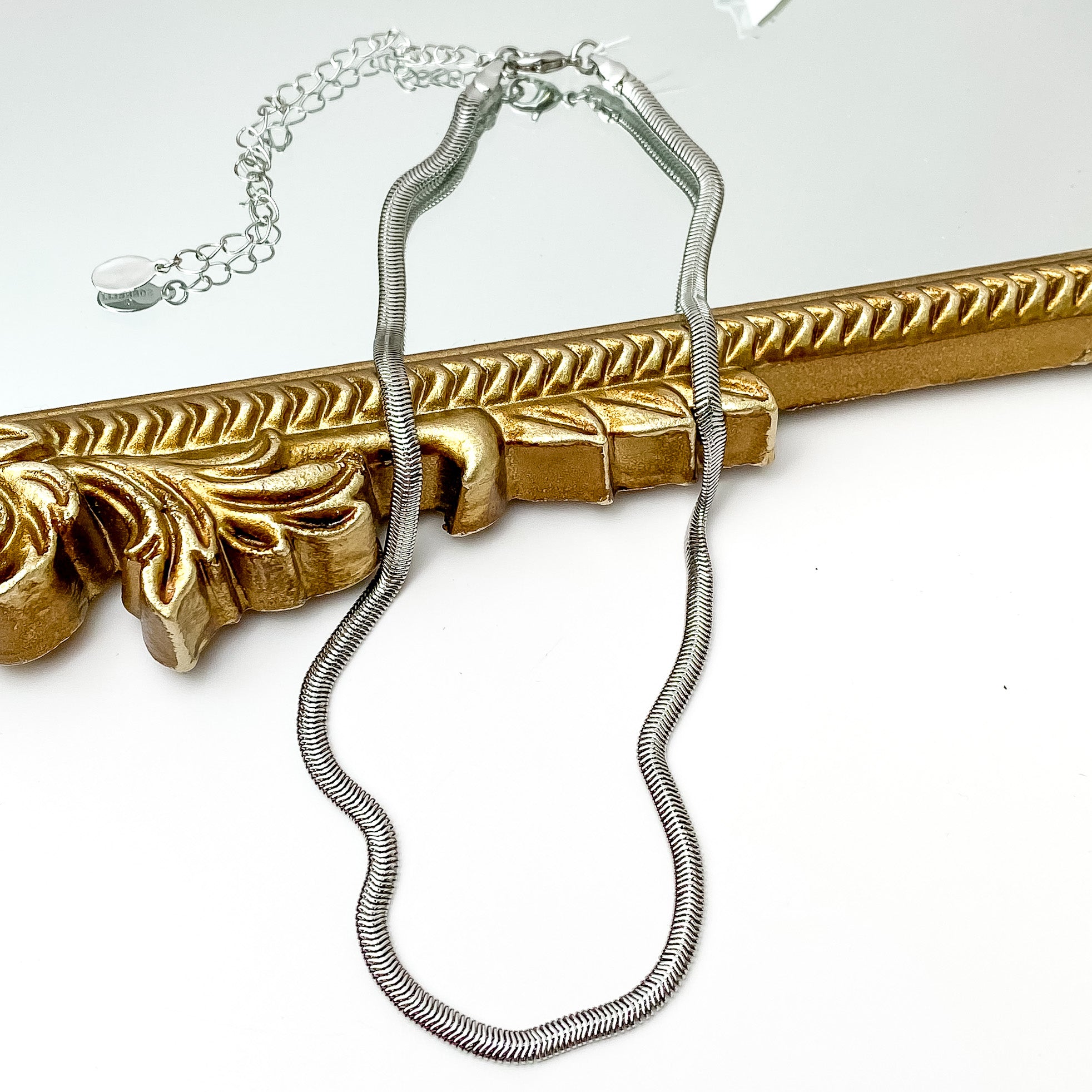 Silver herringbone chain necklace. This necklace is pictured laying partially on a gold mirror on a white background.   