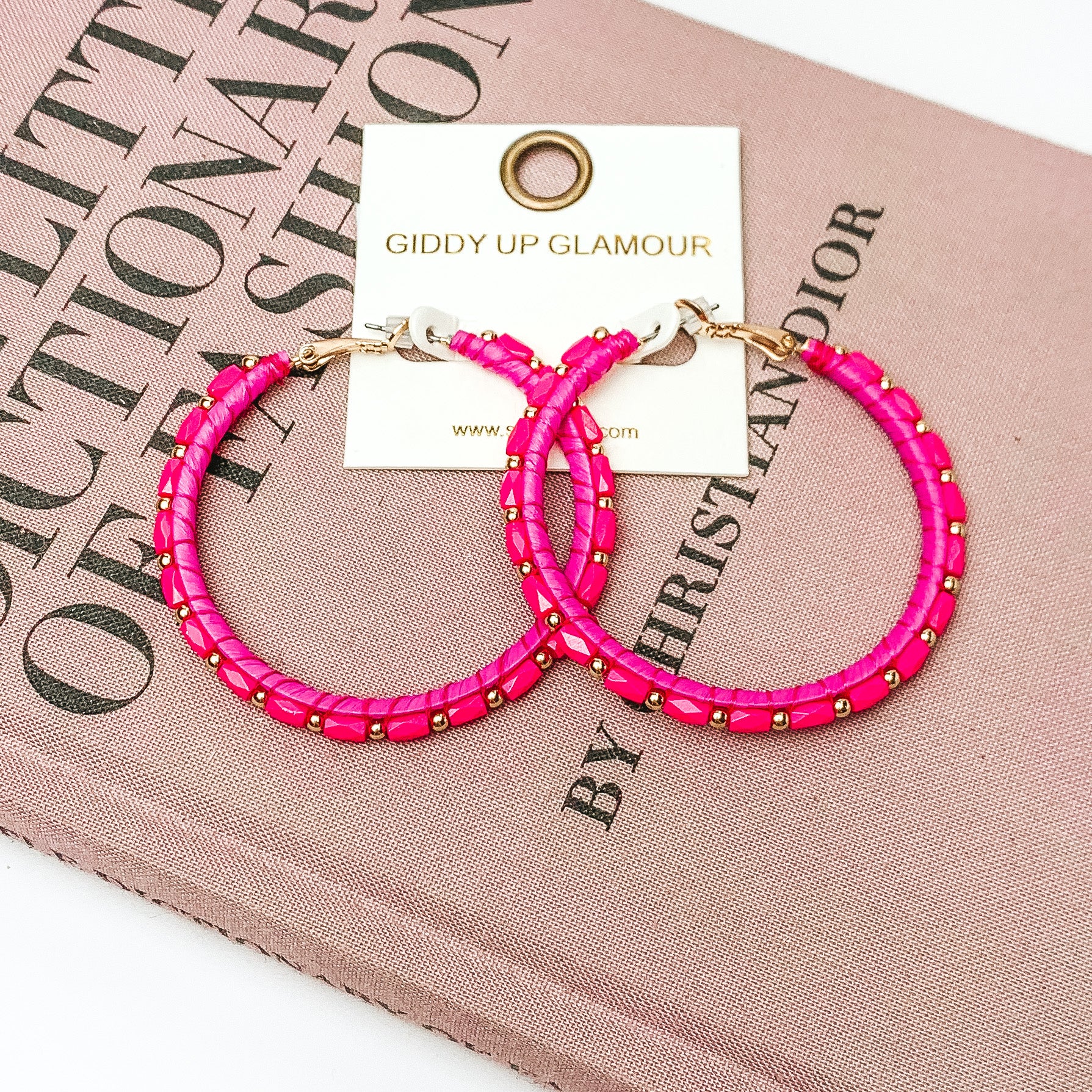 Pictured are circle beaded hoop earrings with gold spacers in hot pink. They are pictured with a pink fashion journal on a white background