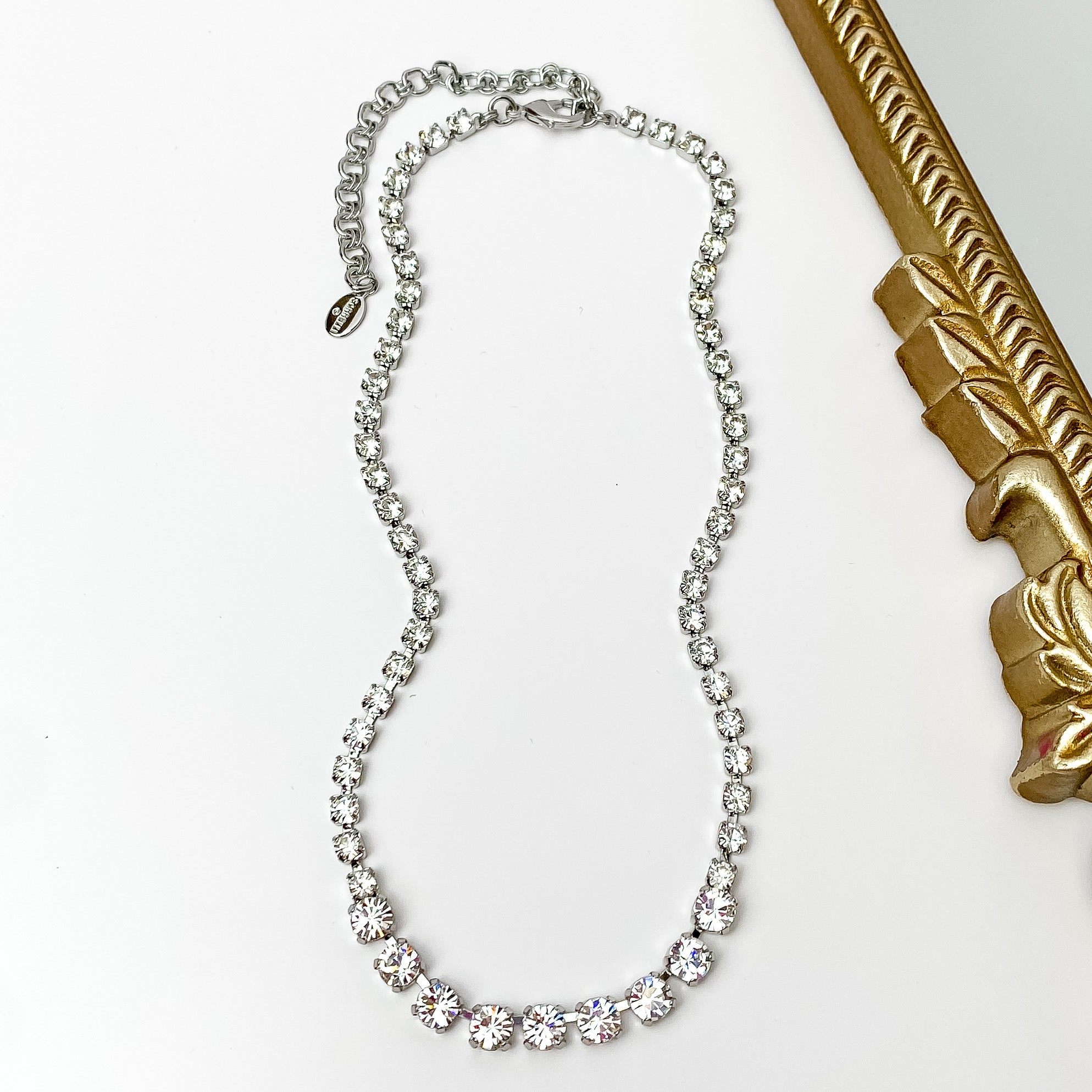 Pictured is a silver necklace with clear crystals. This necklace is pictured on a white background with a gold mirror on the right side.